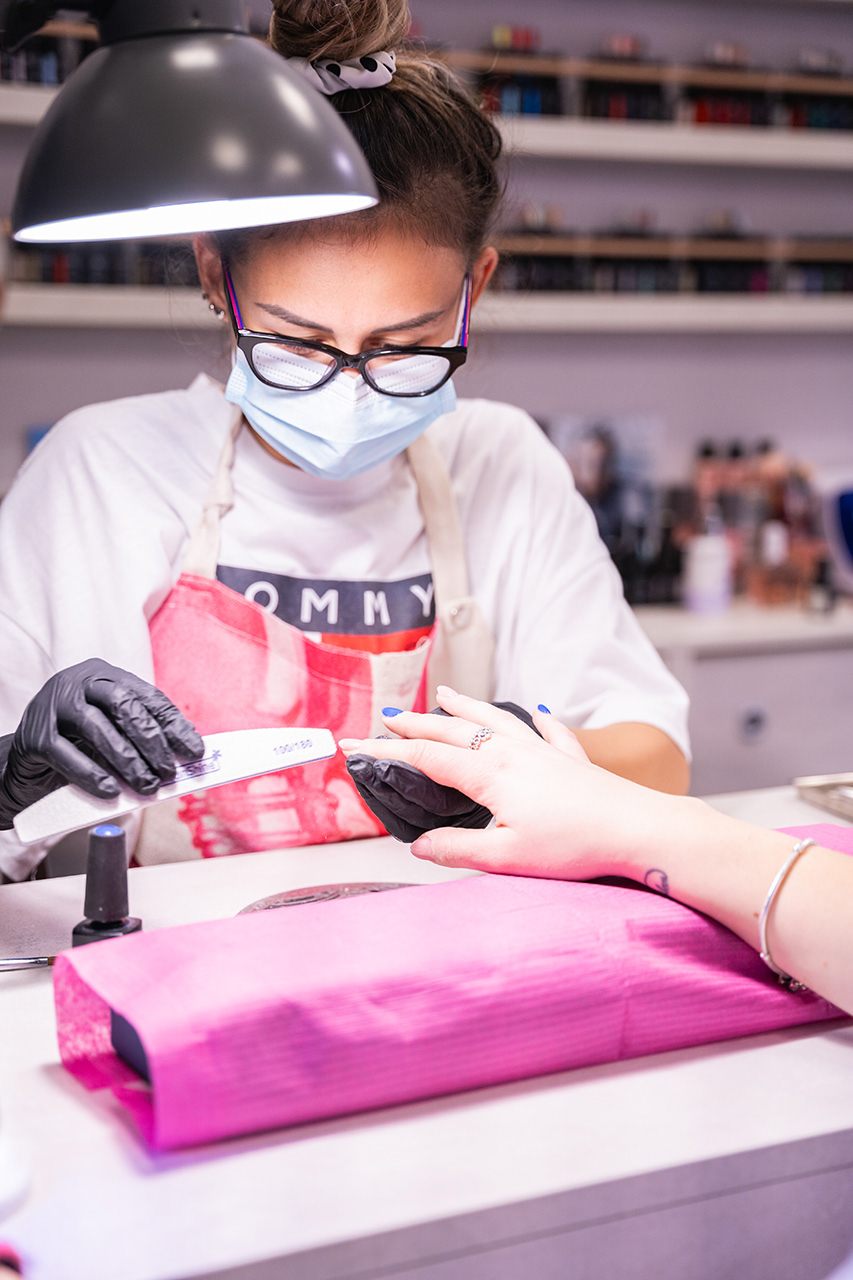 Professional branding photography of a nail technician with a mask and glasses performing a precision manicure at a pink workstation. Capturing authentic beauty salon workflow for marketing use. Specialist retail imagery by Arcane Media.