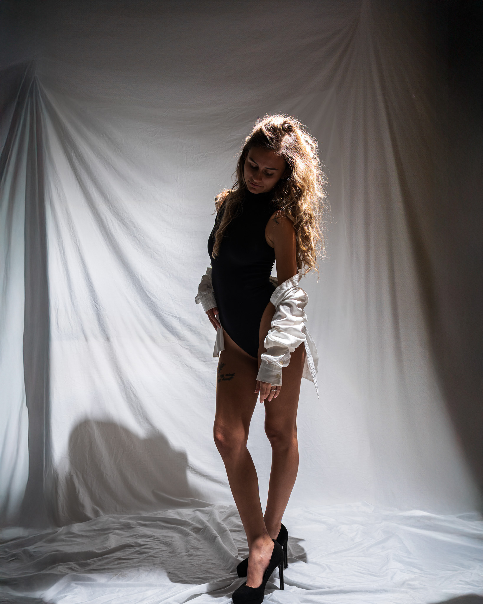 Editorial studio fashion photography featuring a female model in a black bodysuit and white open shirt. The composition utilizes a spotlight technique against a white draped background to create a professional, high-end commercial aesthetic. Storytelling by Arcane Media