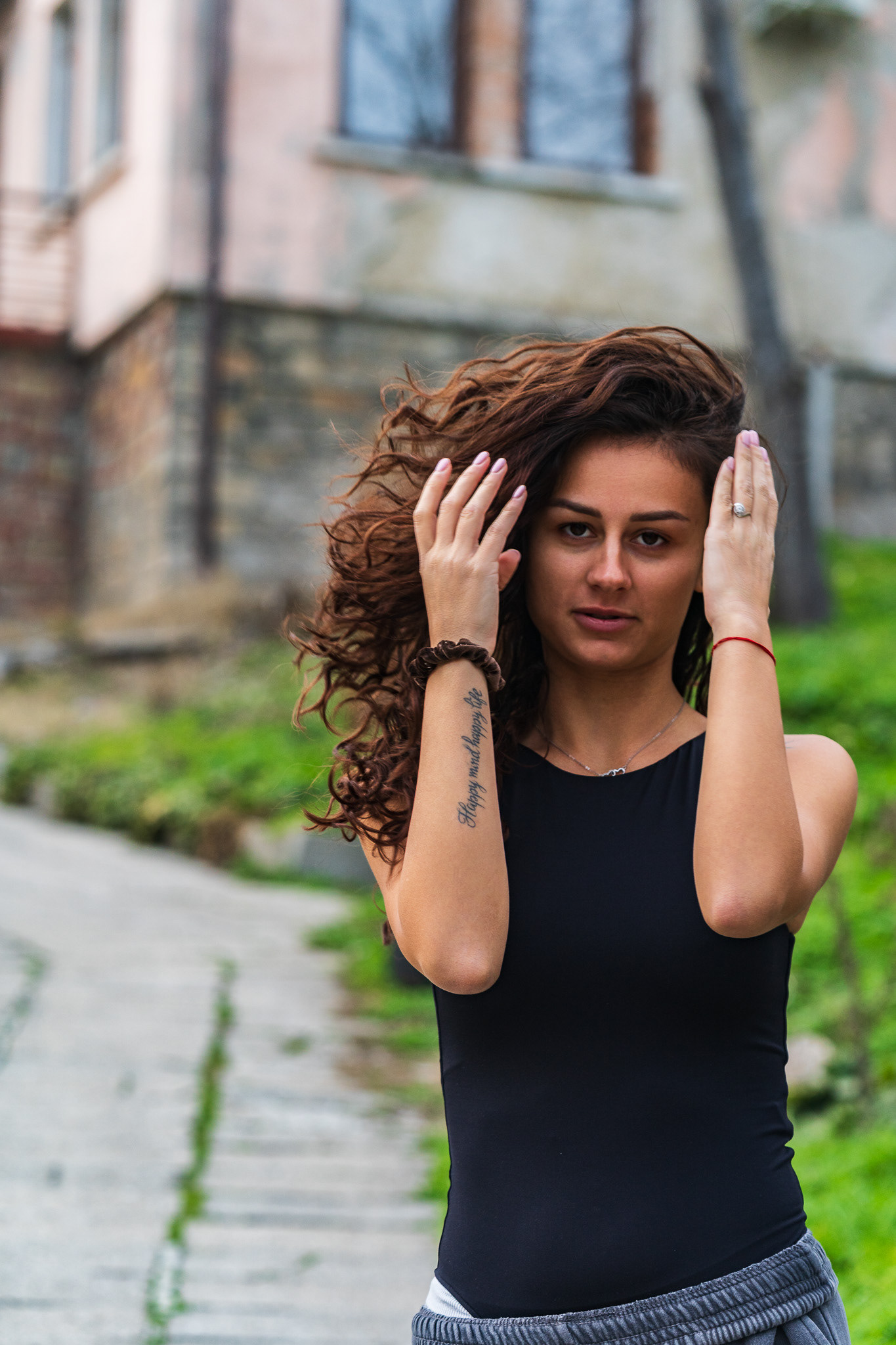 Natural outdoor portrait of a brunette model with curly hair posed against a green hillside and stone building. Editorial photography by Tenyo Barbolski, UK.