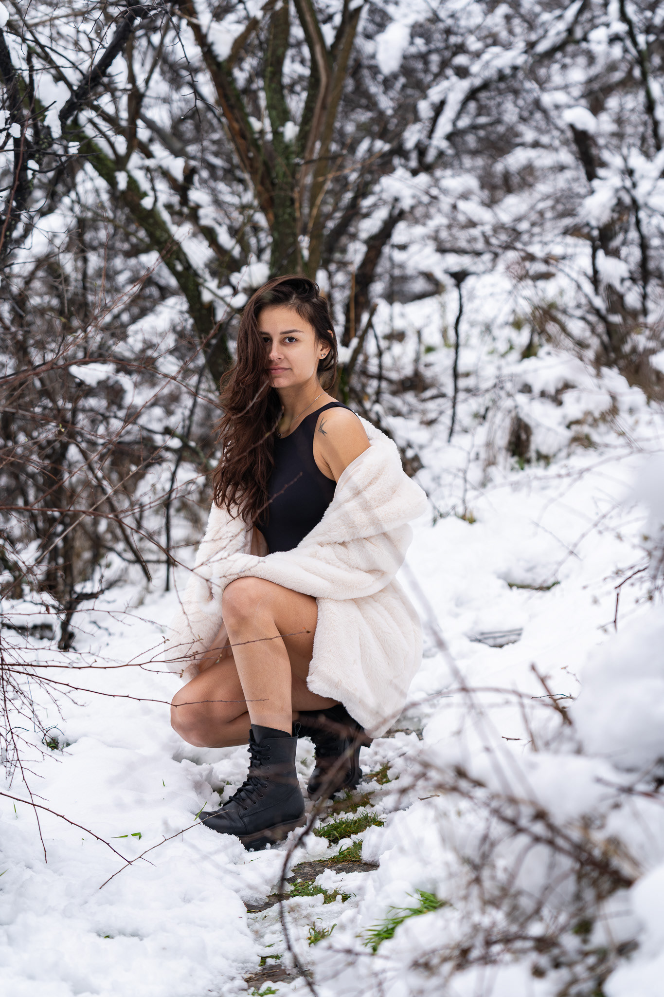 Fashion portrait of a female model in a black bodysuit and white fur coat seated low among snow-covered branches. Cinematic winter photography by Tenyo Barbolski.