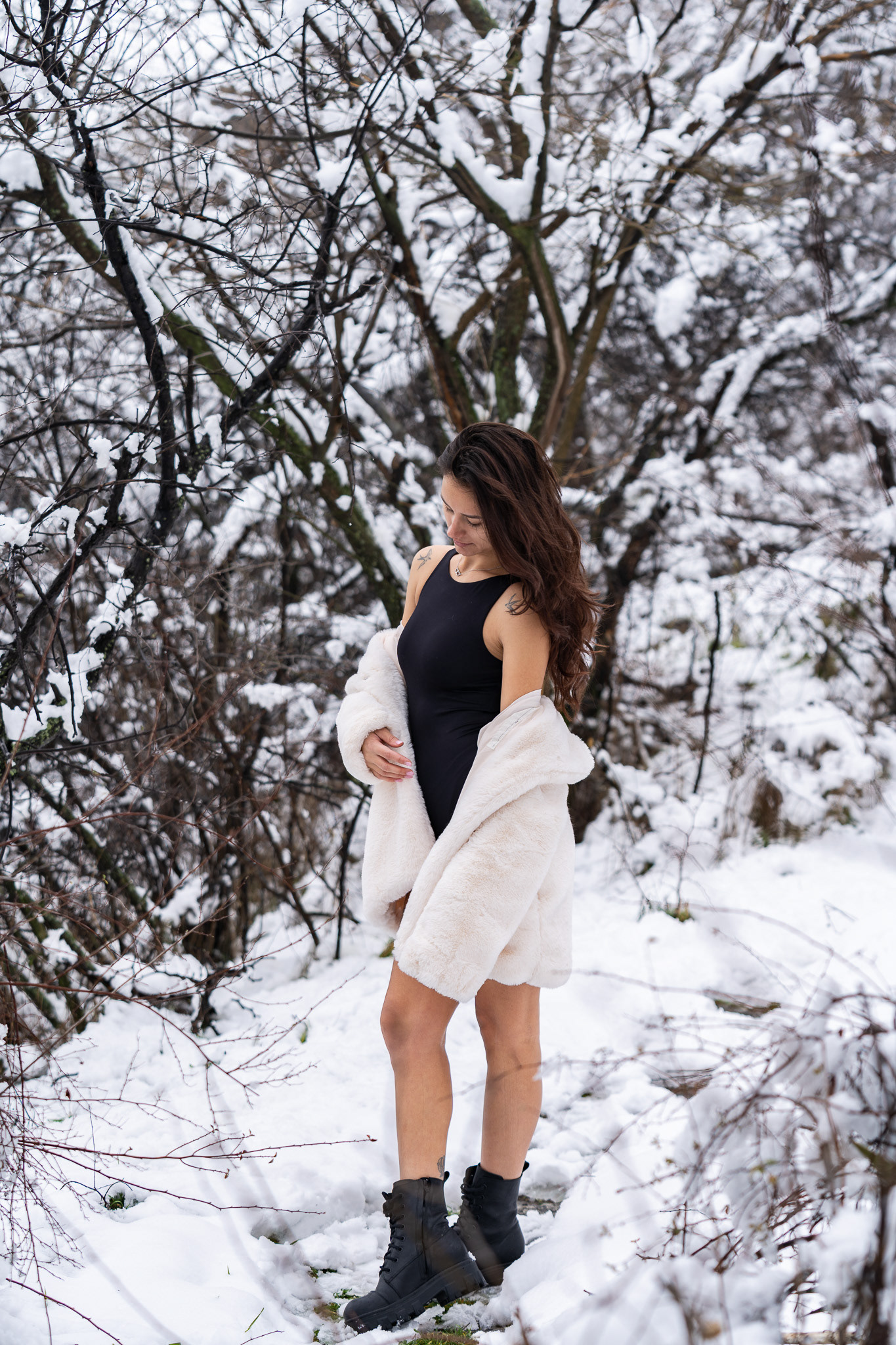 Atmospheric winter fashion portrait of a brunette model in a black bodysuit and white fur coat standing among snow-laden branches. Photography by Tenyo Barbolski, UK.