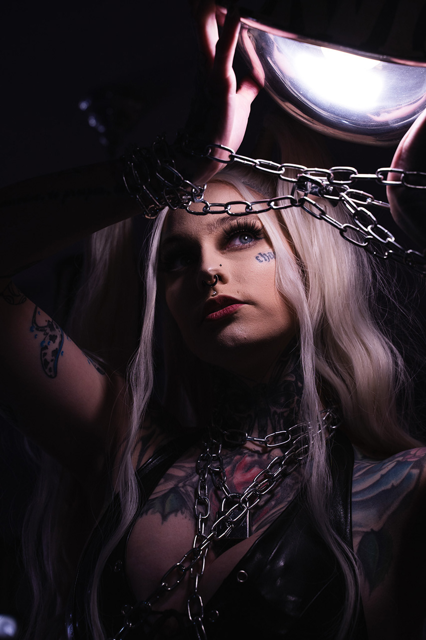 Gritty, high-contrast close-up portrait of a tattooed model featuring chains and low-key lighting. Demonstrating technical proficiency in complex texture and shadow management. Gritty-edgy branding.