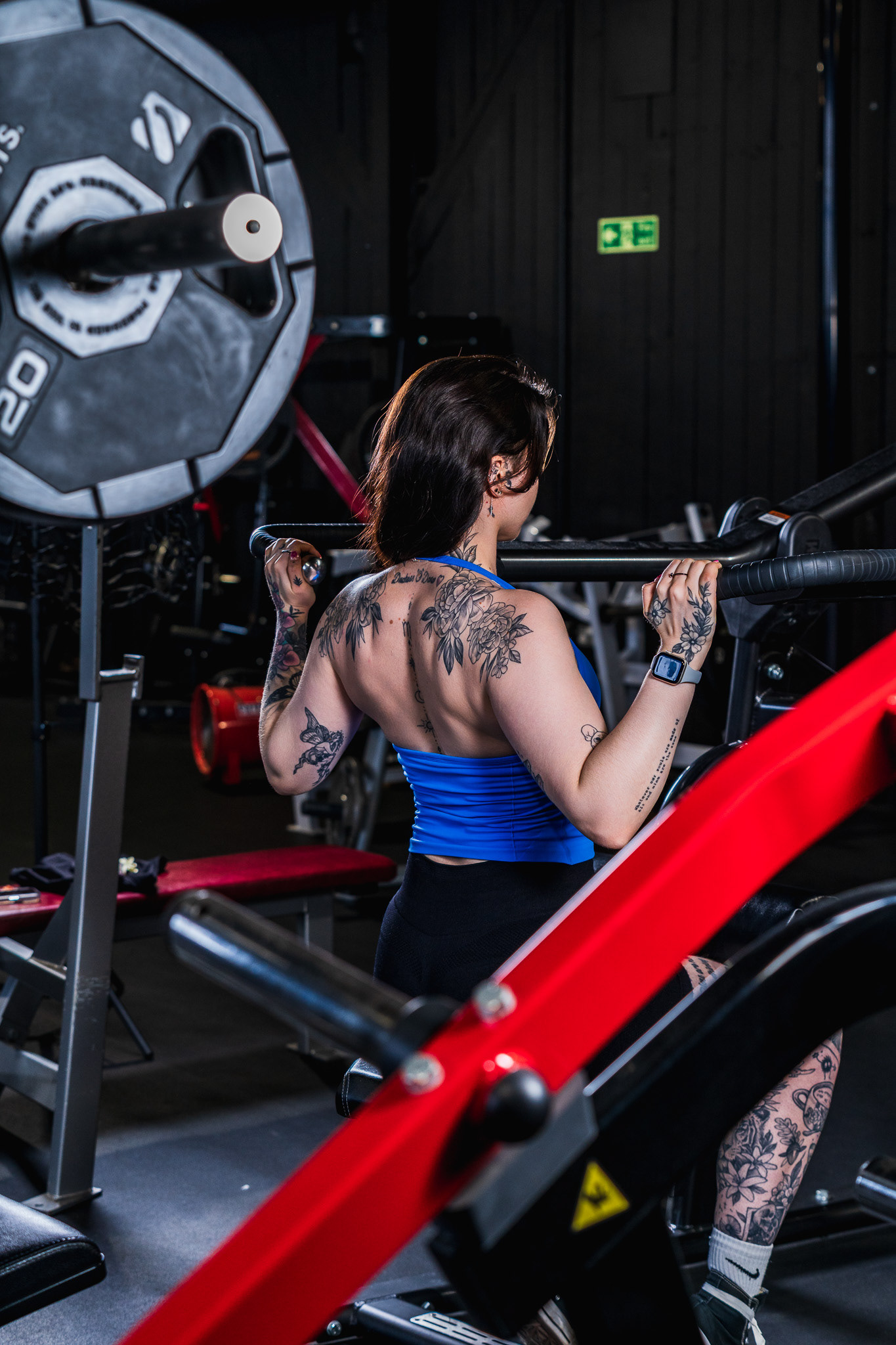 Tattooed female model photographed from behind lifting a barbell overhead in a dark gym. High-contrast editorial fitness photography by Arcane Media, UK.