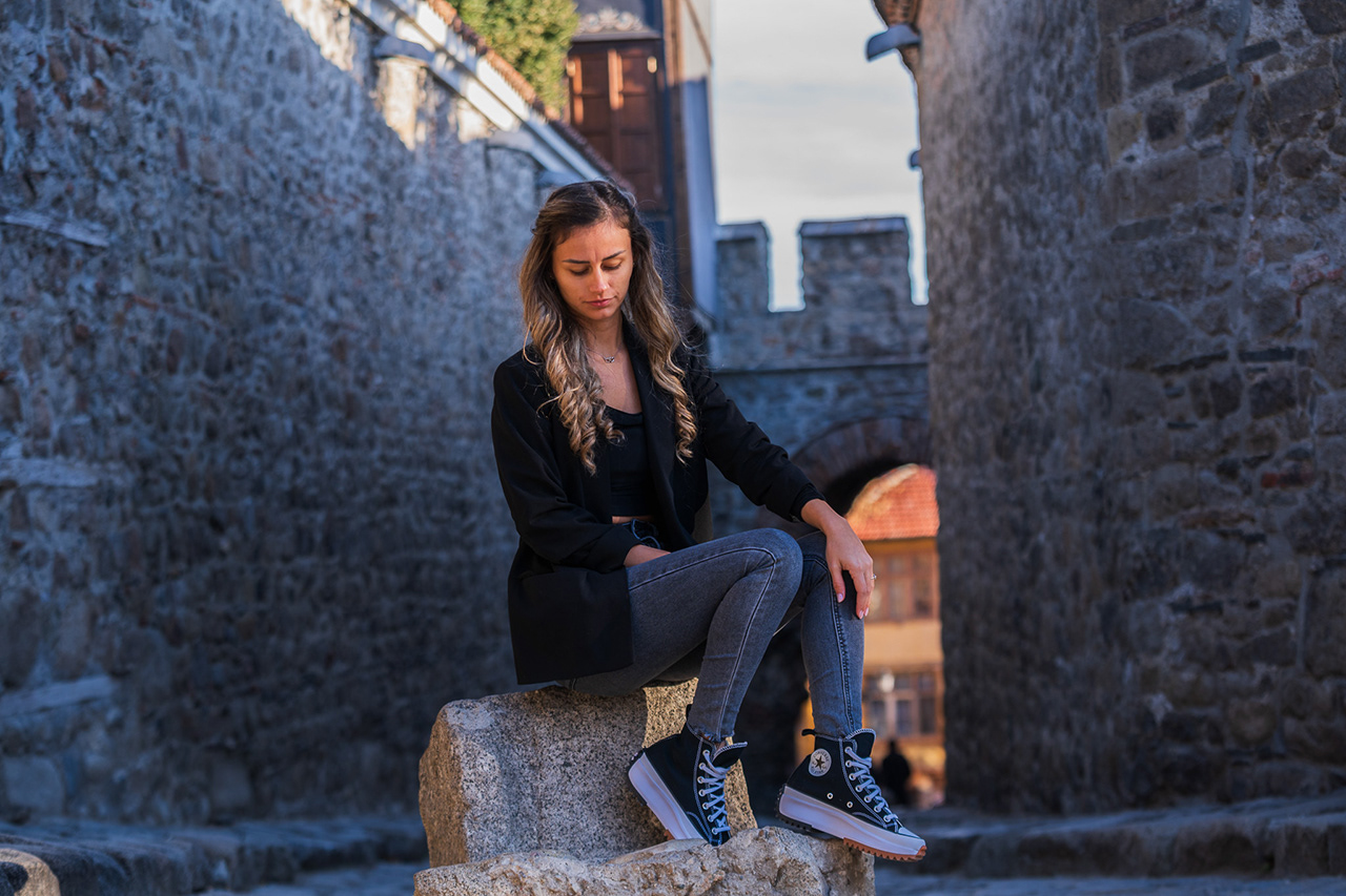 Cinematic fashion portrait of a model seated on a stone block in an ancient corridor. Focused on architectural shadows and timeless lifestyle photography