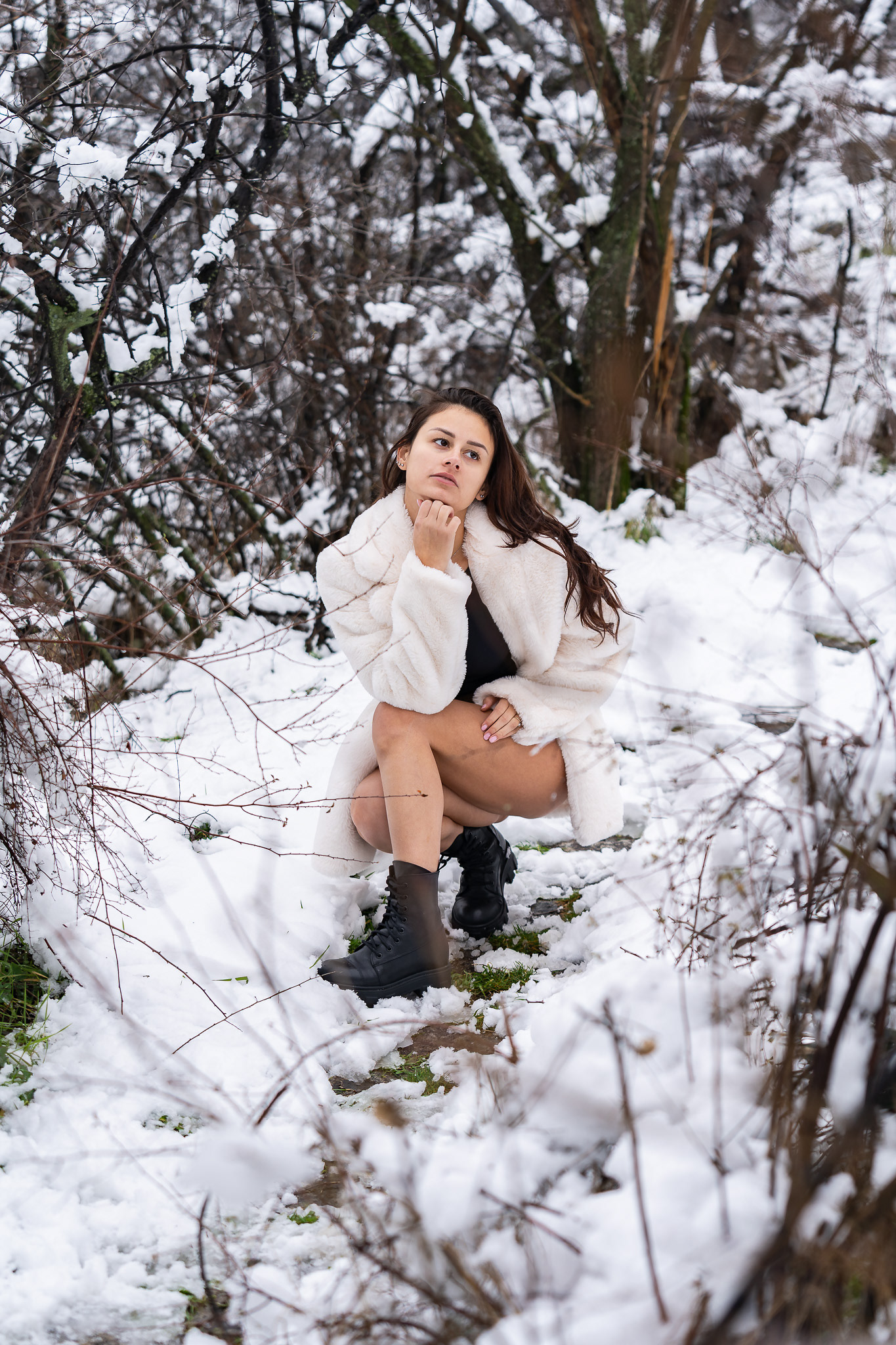 Candid editorial winter portrait of a brunette model in a black bodysuit and white fur coat among snowy trees with a natural relaxed expression. Tenyo Barbolski.