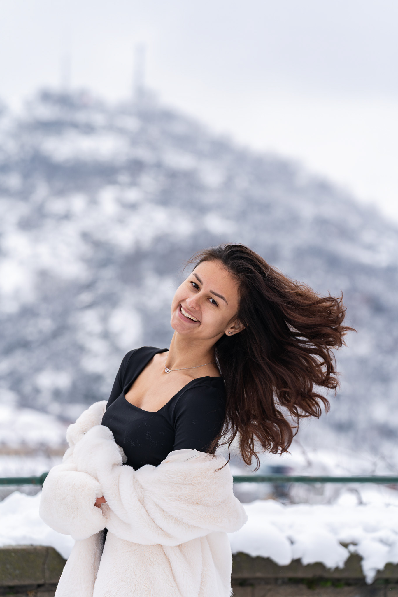 Joyful winter fashion portrait of a model with flowing hair in a white fur coat against a snowy mountain and overcast sky. Editorial photography by Tenyo Barbolski.