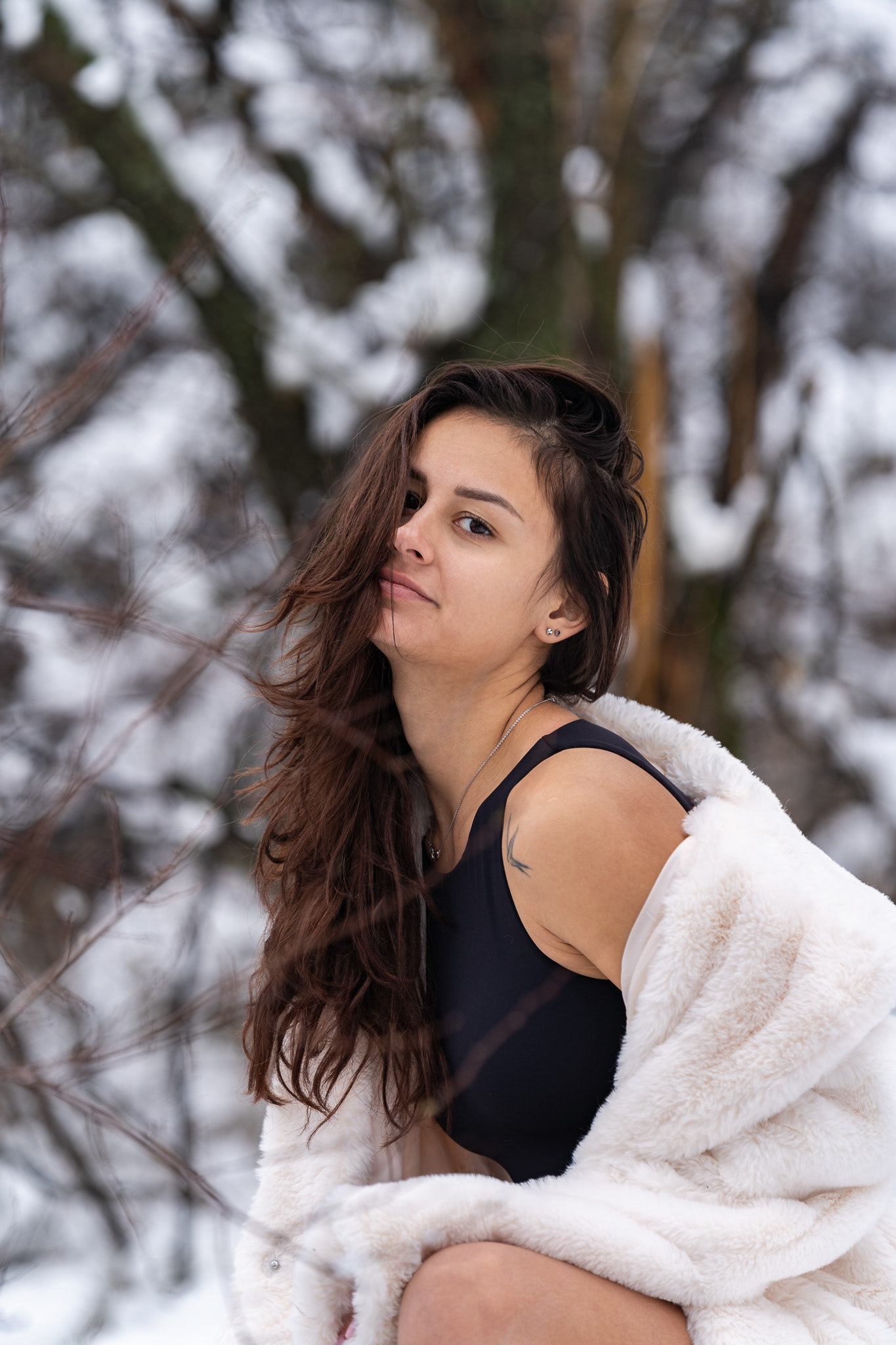 Intimate close-up winter portrait of a brunette model with flowing hair draped in a white fur coat among bare snowy branches. Photography by Tenyo Barbolski.