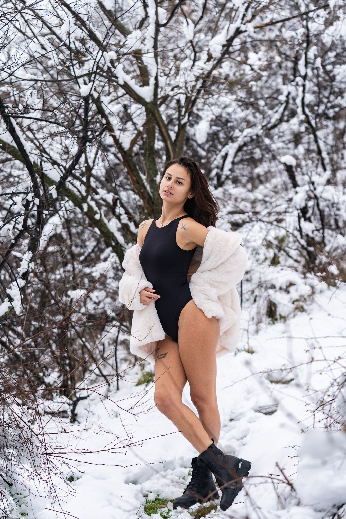 Confident winter fashion portrait of a brunette model in a black bodysuit and white fur coat standing among frost-covered branches. Photography by Arcane Media, UK.