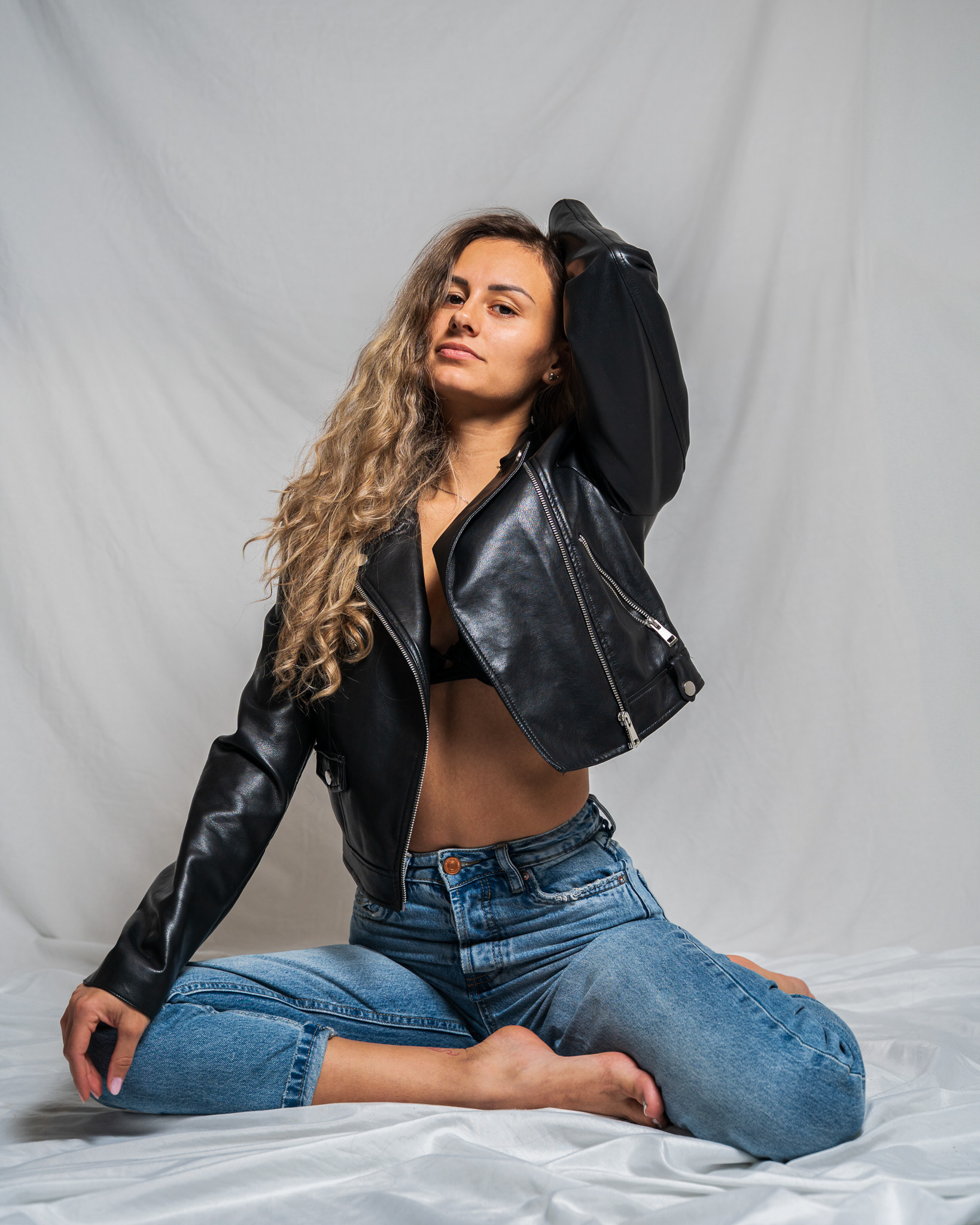 ymmetric studio fashion portrait of a model seated cross-legged in a leather jacket and high-waisted denim. This high-resolution image focuses on central composition and confident gaze for a strong, independent brand identity. Arcane Media.