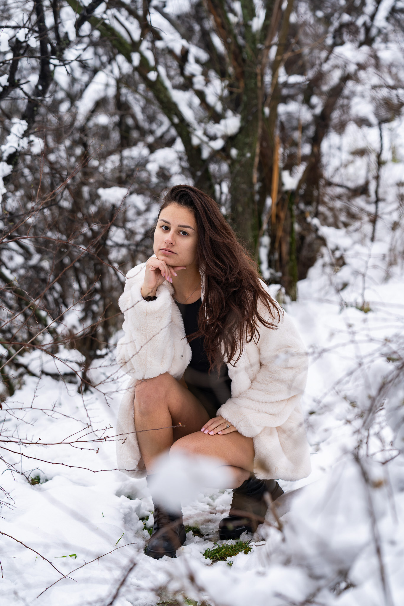 Thoughtful editorial portrait of a brunette model in a black bodysuit and white fur coat crouching in a snowy woodland setting. Photography by Arcane Media, UK.