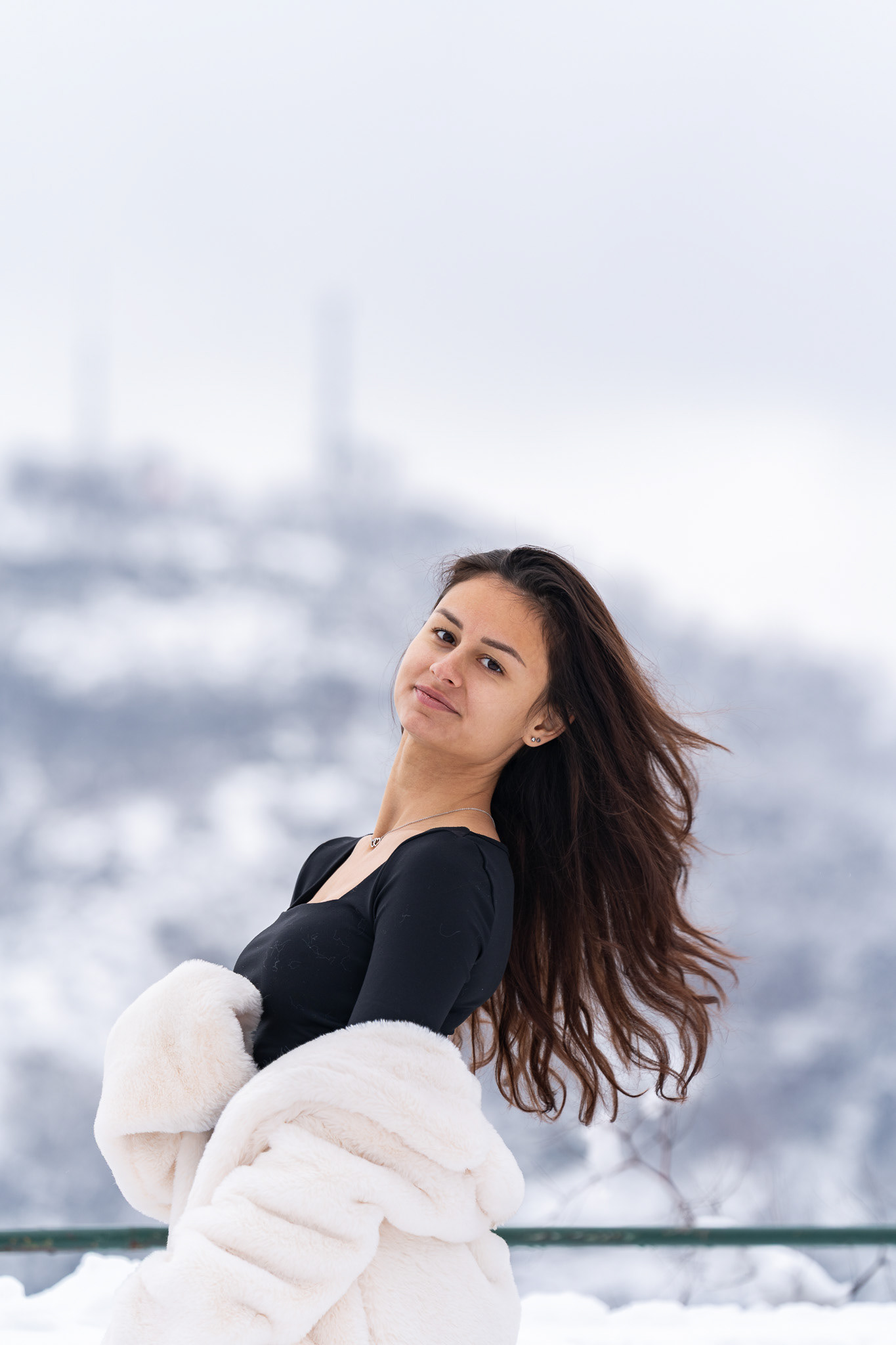 Dynamic close-up winter portrait of a brunette model with flowing hair in a black top and white fur coat against a snowy mountain skyline. Arcane Media, UK.