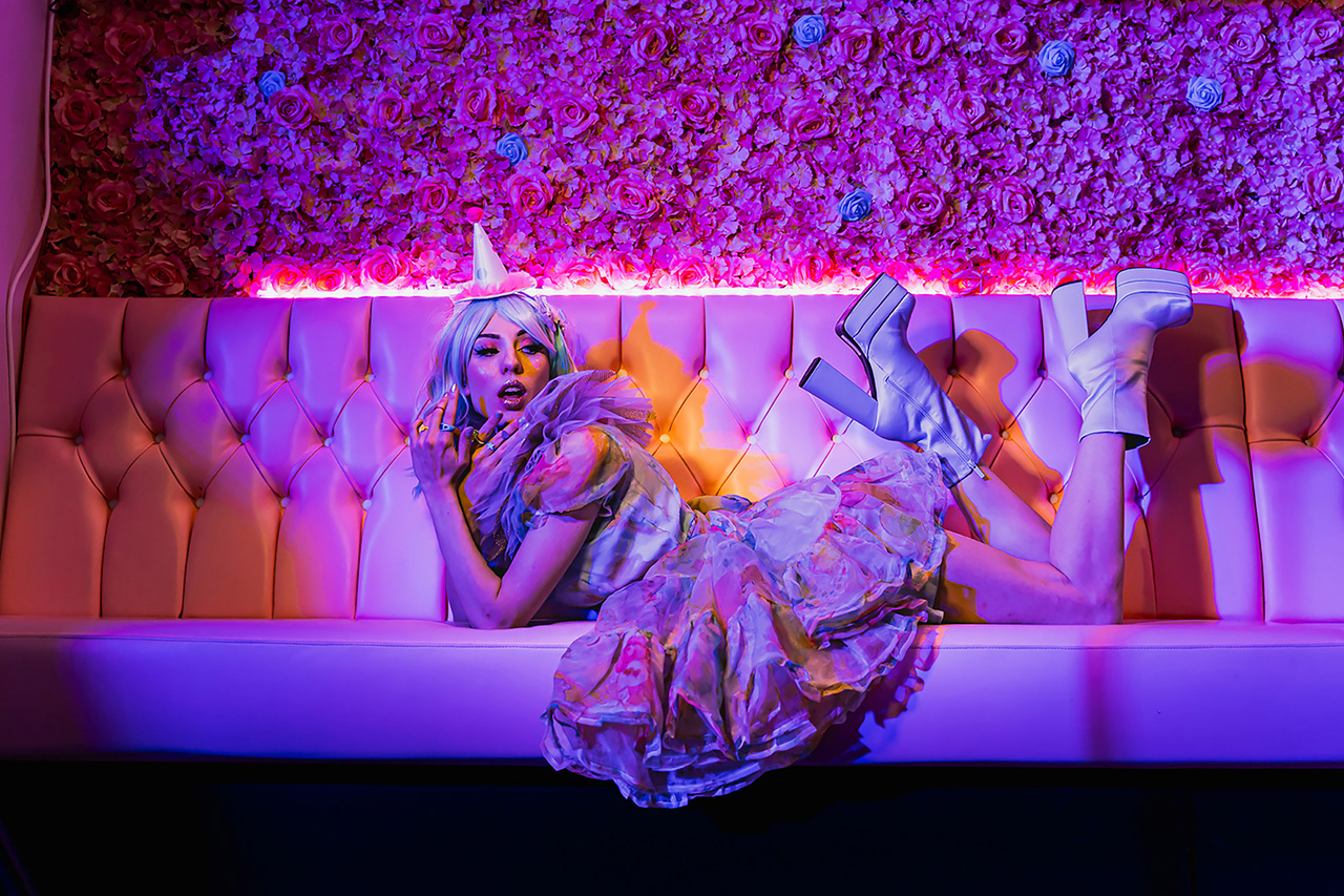 Conceptual 'Pastel Goth' fashion editorial. Neon-lit portrait of a model with pastel purple hair and a unicorn horn, posing with dramatic low-key lighting for a mysterious, ethereal mood