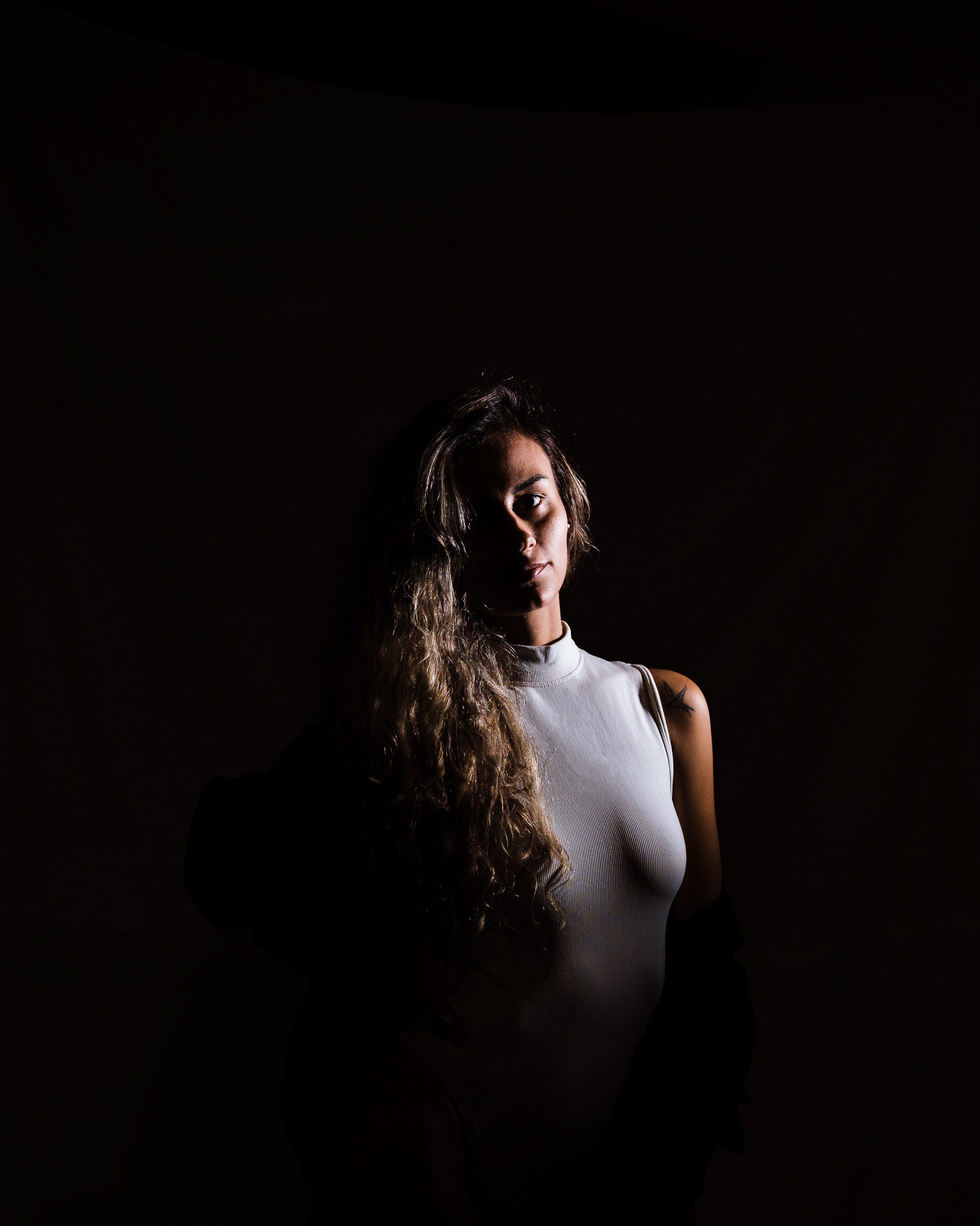 Dramatic low-key portrait of a model with long wavy hair. This technical shot focuses on chiaroscuro lighting, where deep shadows isolate the subject for a mysterious and intense visual narrative. Fine art portraiture by Tenyo Barbolski.