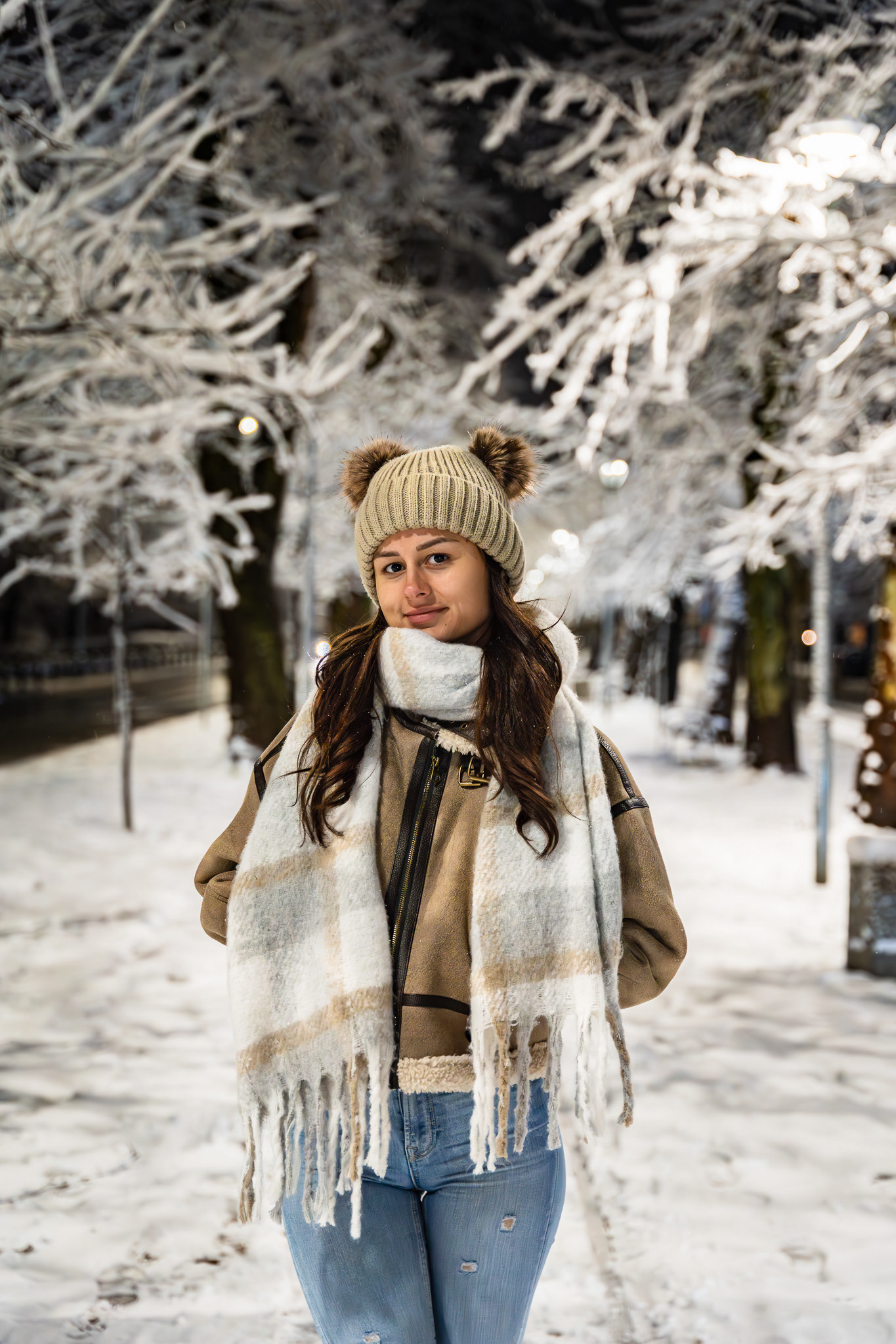 Evening winter fashion portrait of a female model in a tan shearling coat, beanie and scarf among snow-covered trees at night. Editorial photography by Tenyo Barbolski.