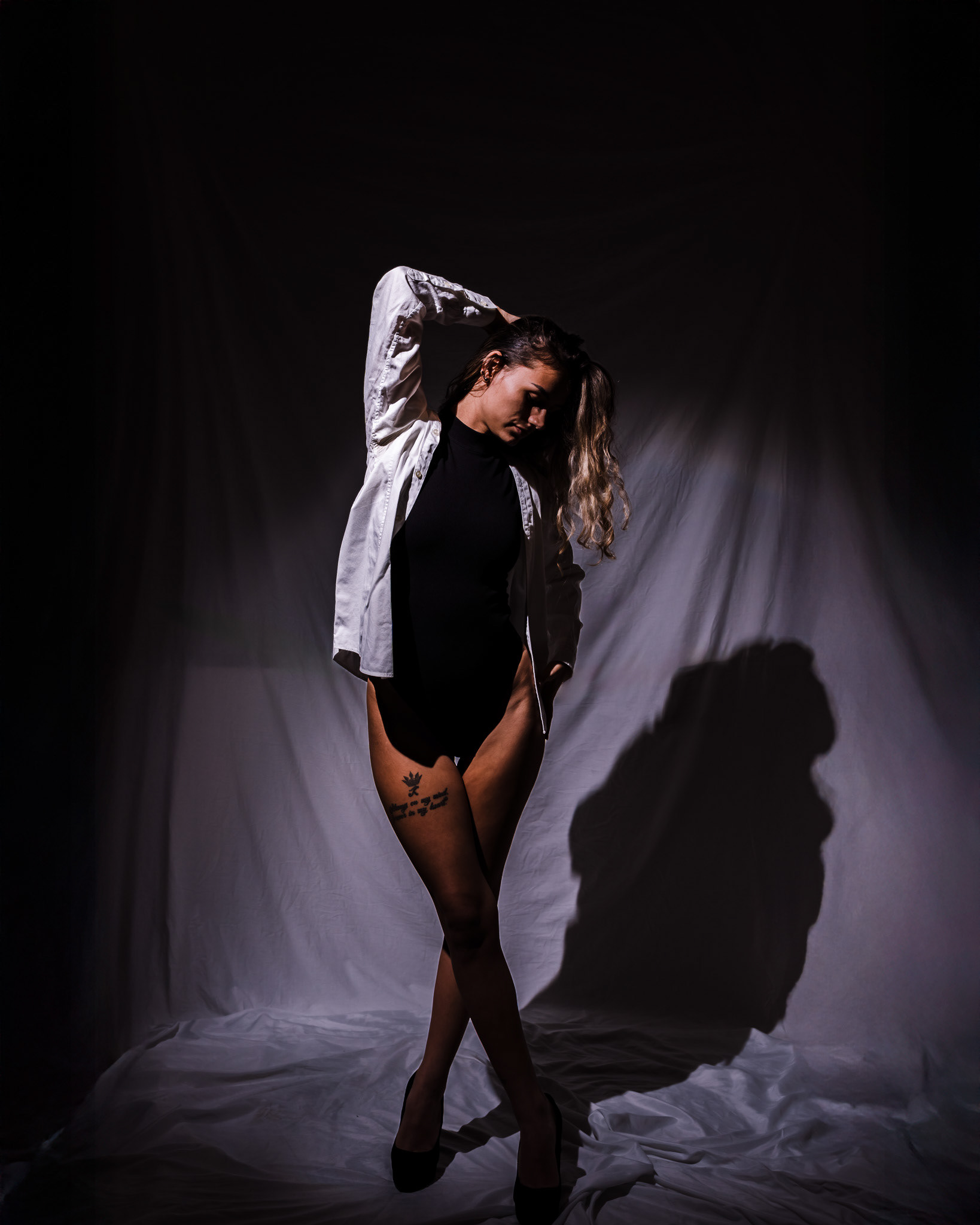 A dramatic, conceptual studio portrait of a model utilizing dramatic, low-key lighting and strong shadows. Focused on the relationship between the body, clothing textures (white shirt/black bodysuit), and deep background fabric to create a profound and mysterious visual narrative