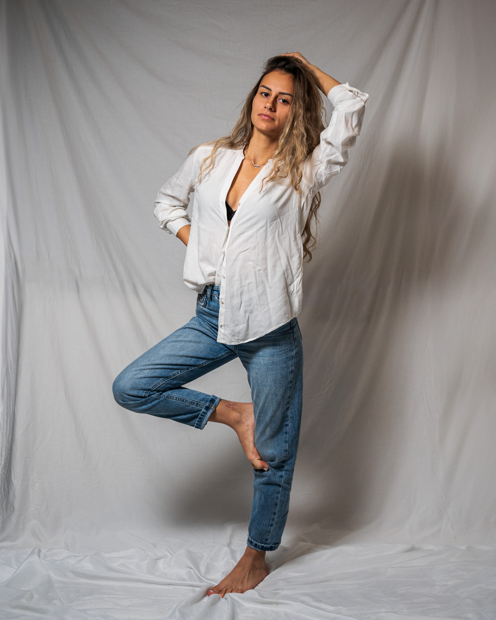Lifestyle-focused portrait of a model in blue jeans and a white linen shirt performing a balanced tree pose. Captured against a minimalist studio background to highlight relaxed, natural movement and organic branding. Commercial imagery by Arcane Media