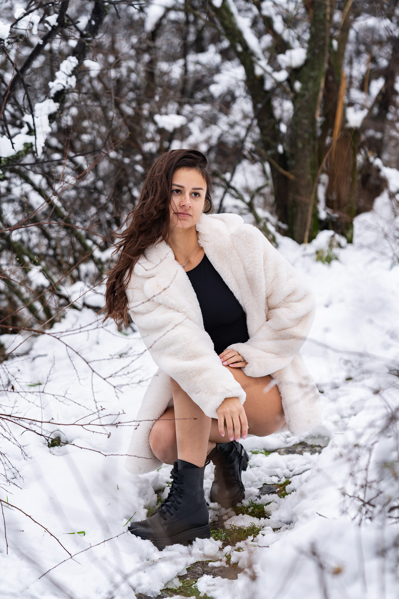 Editorial winter portrait of a brunette model in a black bodysuit and white fur coat seated on a snowy hillside among bare trees. Arcane Media, Cambridgeshire.