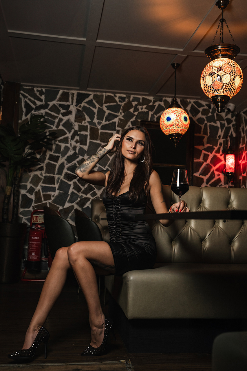 Wide-angle lifestyle shot of a model in a dark dress seated at a bar, showcasing the full interior design and upscale environment.
