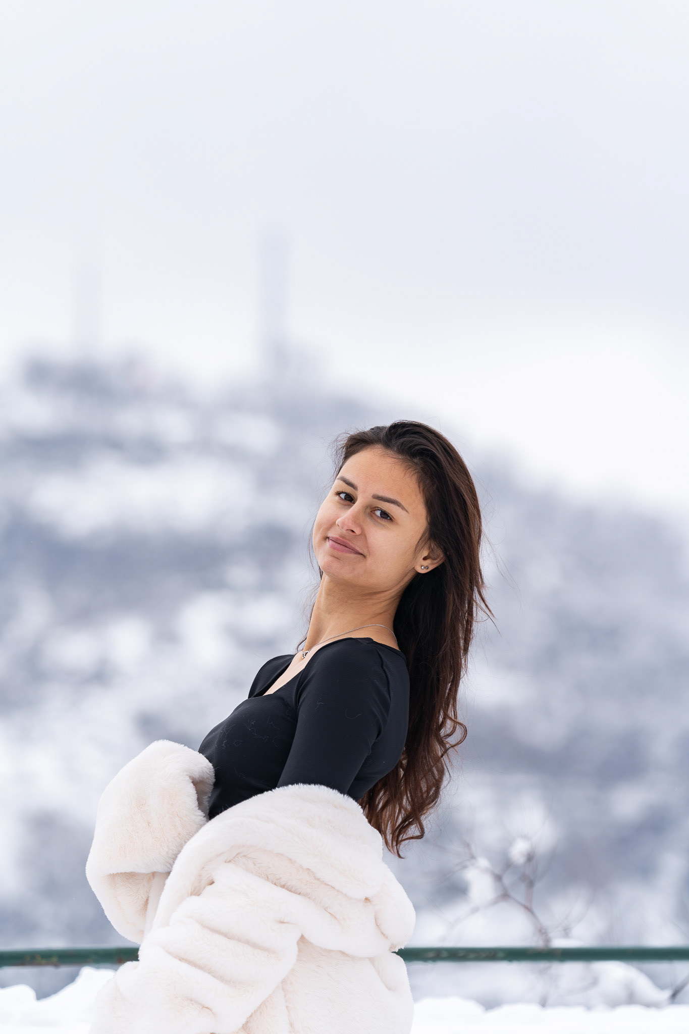Intimate close-up winter portrait of a brunette model in a black top with a white fur coat against a snowy cityscape backdrop. Photography by Arcane Media, UK.