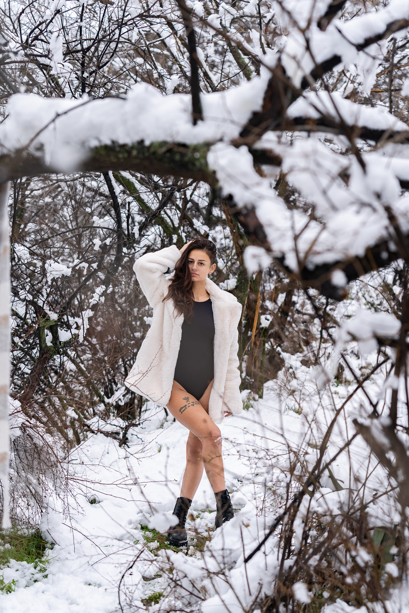 Winter fashion portrait of a brunette model in a white fur coat and black bodysuit among snow-covered trees. Outdoor lifestyle photography by Arcane Media, UK.