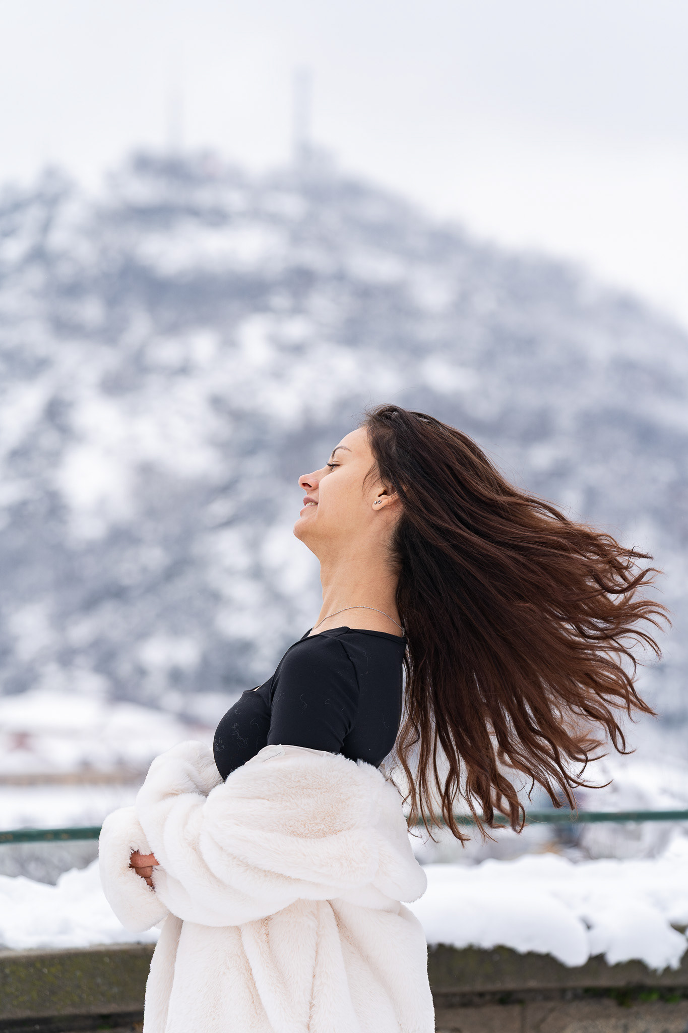 Cinematic winter portrait of a brunette model with wind-swept hair in a black bodysuit and white fur coat with a snow-covered hill behind her. Arcane Media, UK.