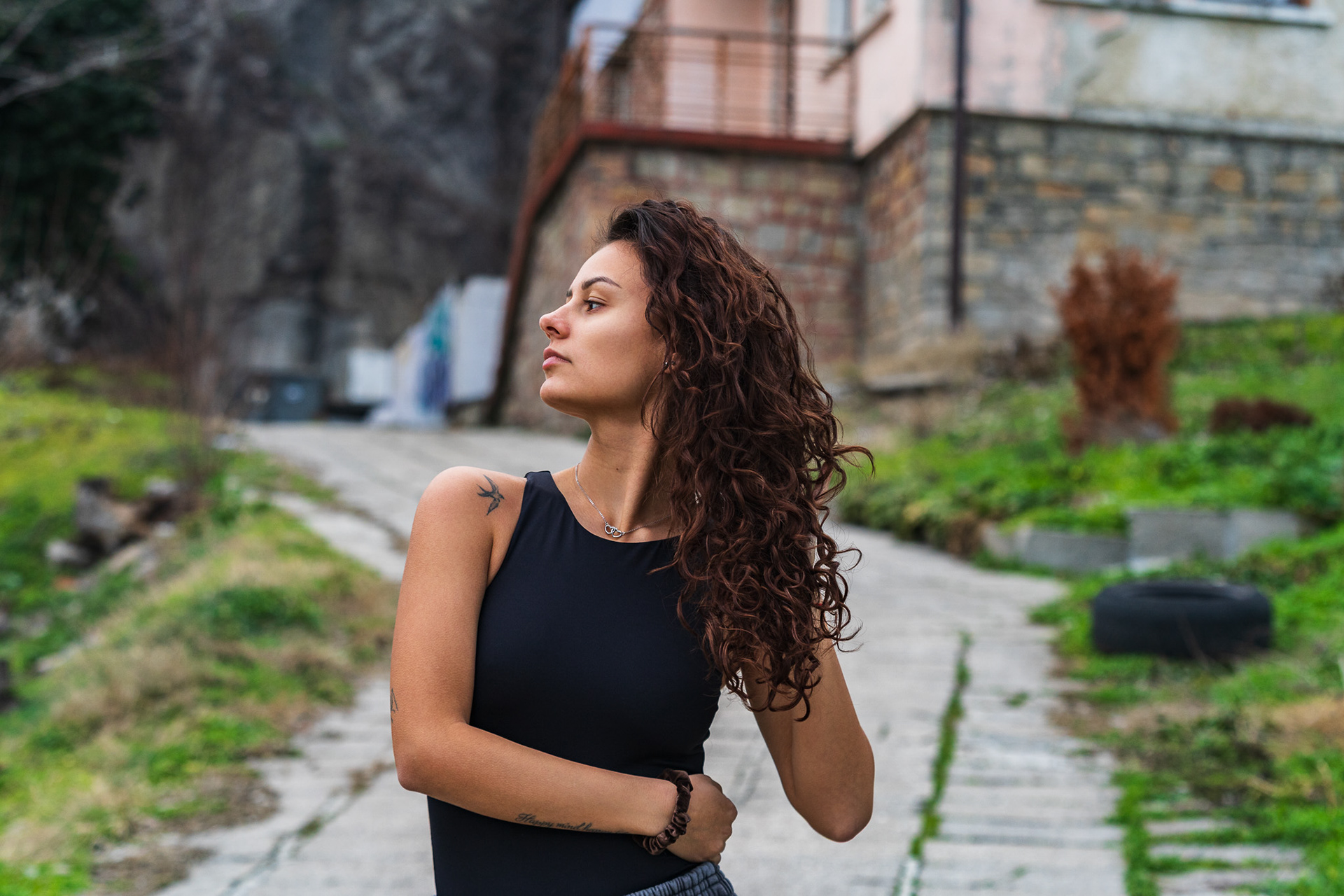 Atmospheric lifestyle portrait of a model looking upward against a blurred stone pathway and lush green surroundings. Editorial photography by Tenyo Barbolski, UK.