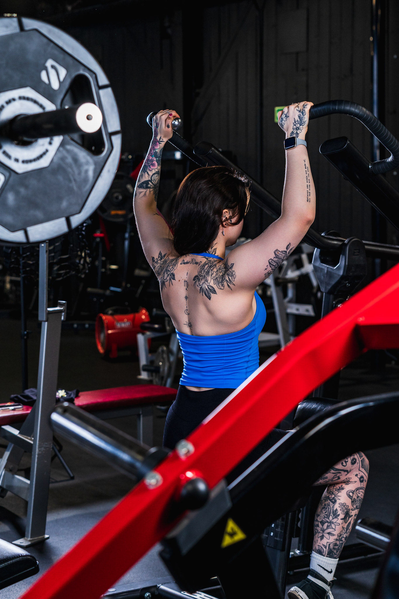 Dramatic overhead barbell shot of a tattooed female model in a gym. Editorial back and physique photography with red equipment contrast by Tenyo Barbolski, Cambridgeshire.
