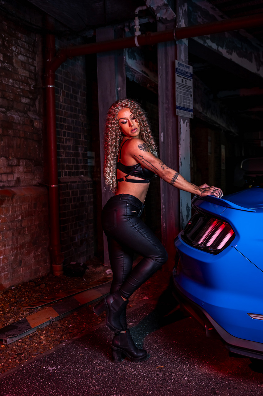 Dynamic low-angle portrait of a professional fitness model on the hood of a blue Mustang. Technical retouching balances the sharp highlights of the car with the model’s athletic texture, creating a high-impact commercial 'Hero' shot.