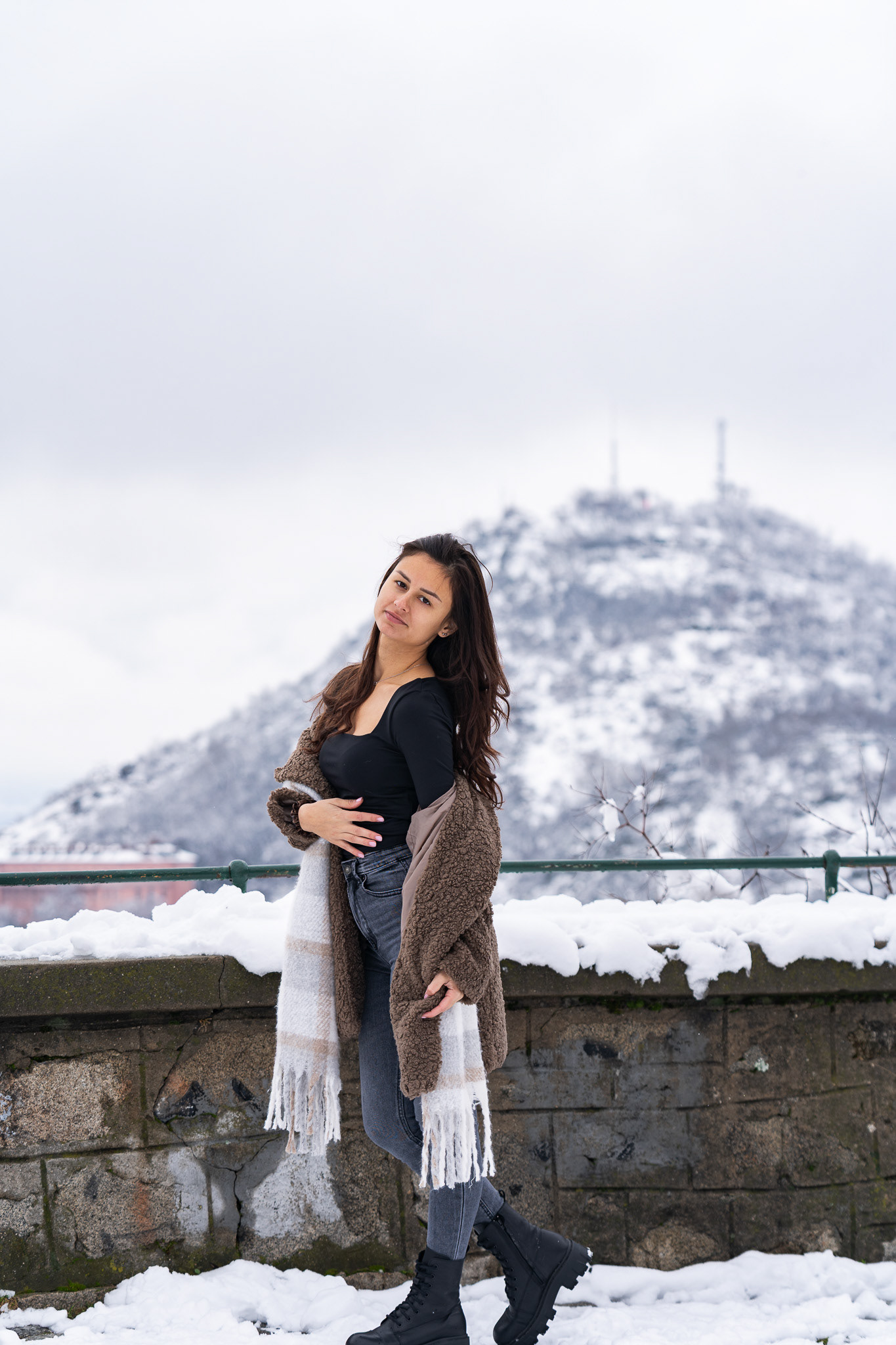 Editorial winter lifestyle portrait of a brunette model in a brown scarf and coat posed against a snow-covered mountain and stone wall. Photography by Tenyo Barbolski.