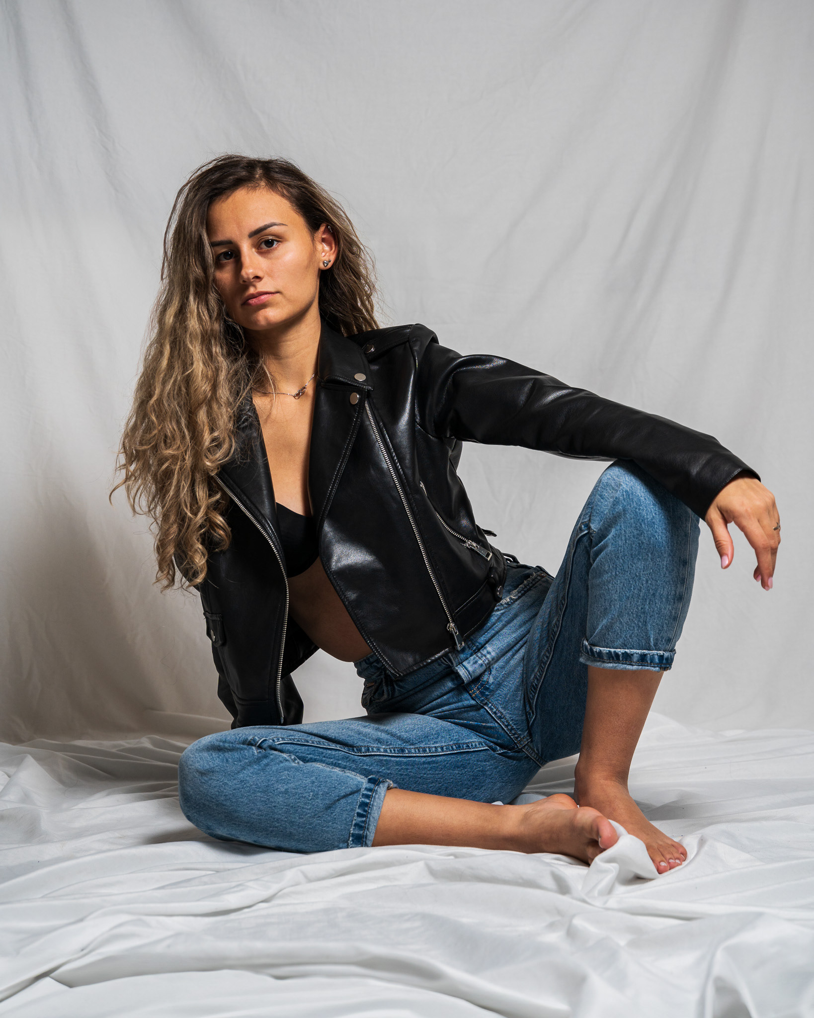 Professional fashion portrait of a model seated in blue jeans and a black leather jacket. Expert lighting emphasizes the contrast between denim and leather textures, suitable for apparel marketing and editorial portfolios. Shot by Tenyo Barbolski.