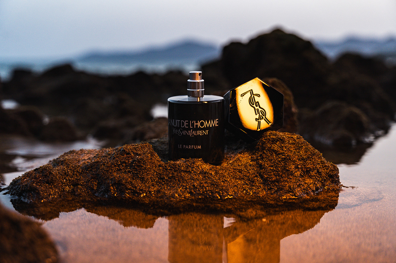 Cinematic commercial product portrait of a YSL perfume bottle and box on wet rocks, using advanced 'through-the-object' lighting to highlight the 'Y' logo. A masterclass in creative commercial composition and detail emphasis."