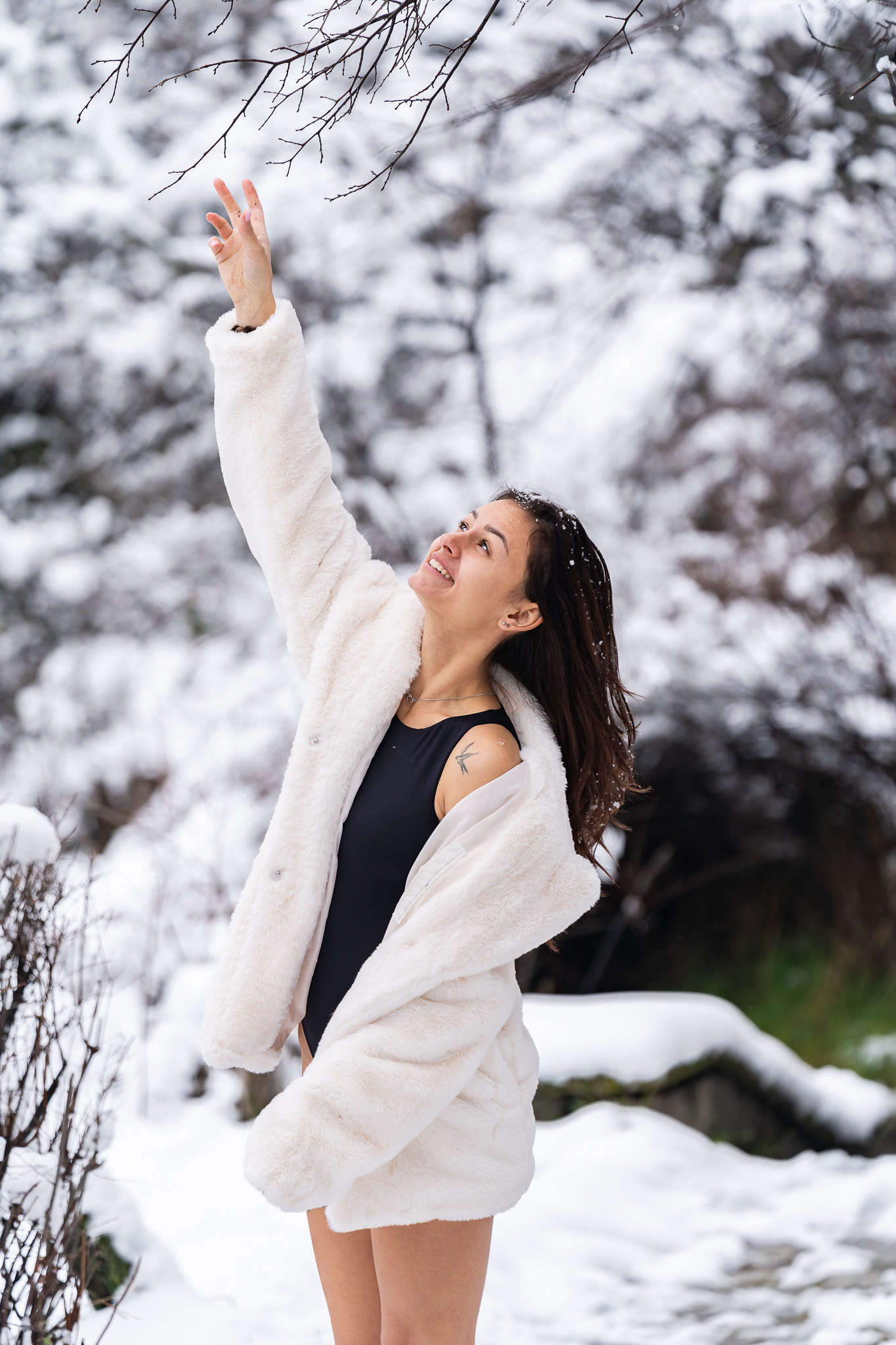 Dynamic winter fashion photography of a brunette model in a white fur coat with arms raised among snow-covered branches. Editorial lifestyle photography by Arcane Media.
