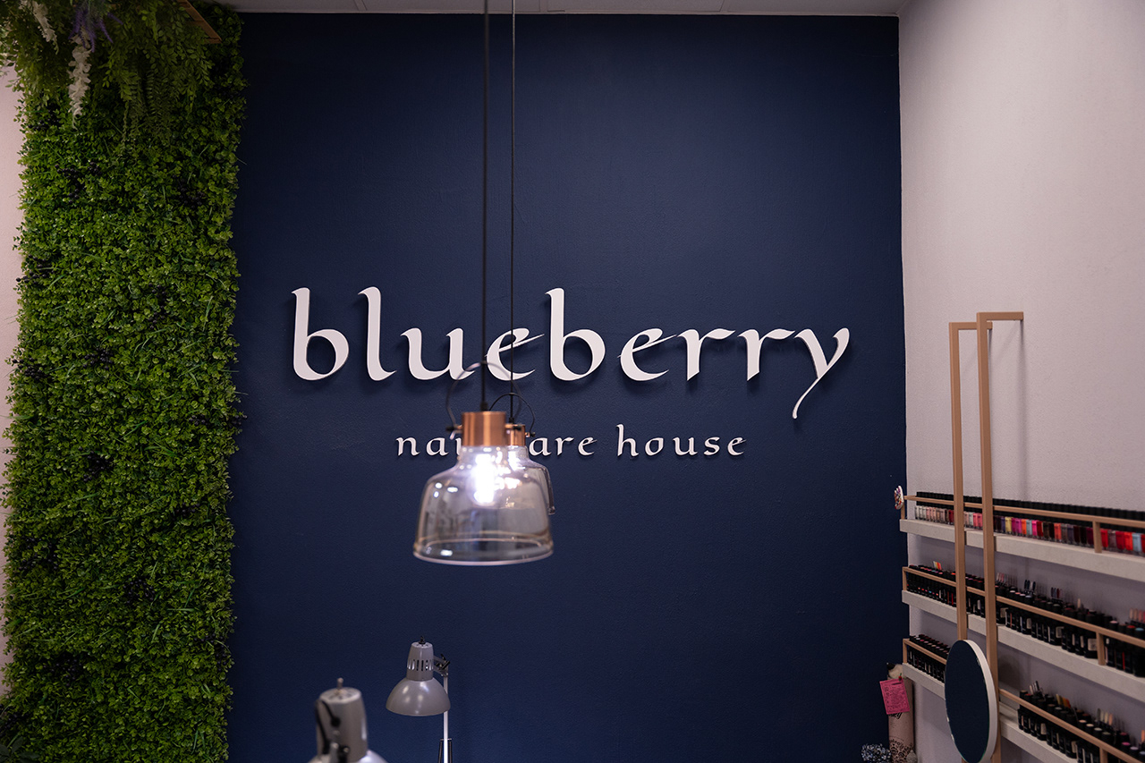 Symmetric commercial interior shot of the 'Blueberry Nail Care House' branding on a deep navy wall, flanked by greenery. Highlighting professional salon logo design and lighting