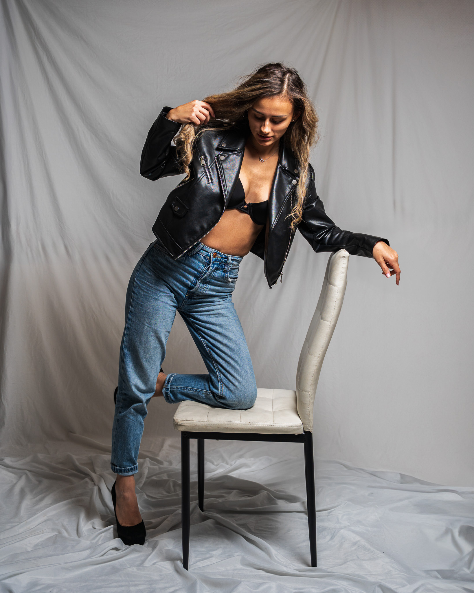 Dynamic editorial fashion shot featuring a model in a leather jacket kneeling on a white studio chair. This composition showcases creative posing and high-fidelity texture detail for a luxury fashion brand identity.