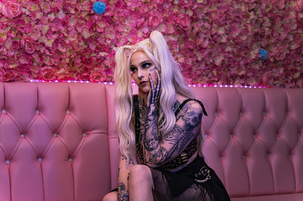 Detailed editorial beauty portrait of a blonde model with extensive tattoos, framed by a neon-pink floral wall and booth. Precise color grading and texture enhancement. Work by Tenyo Barbolski.