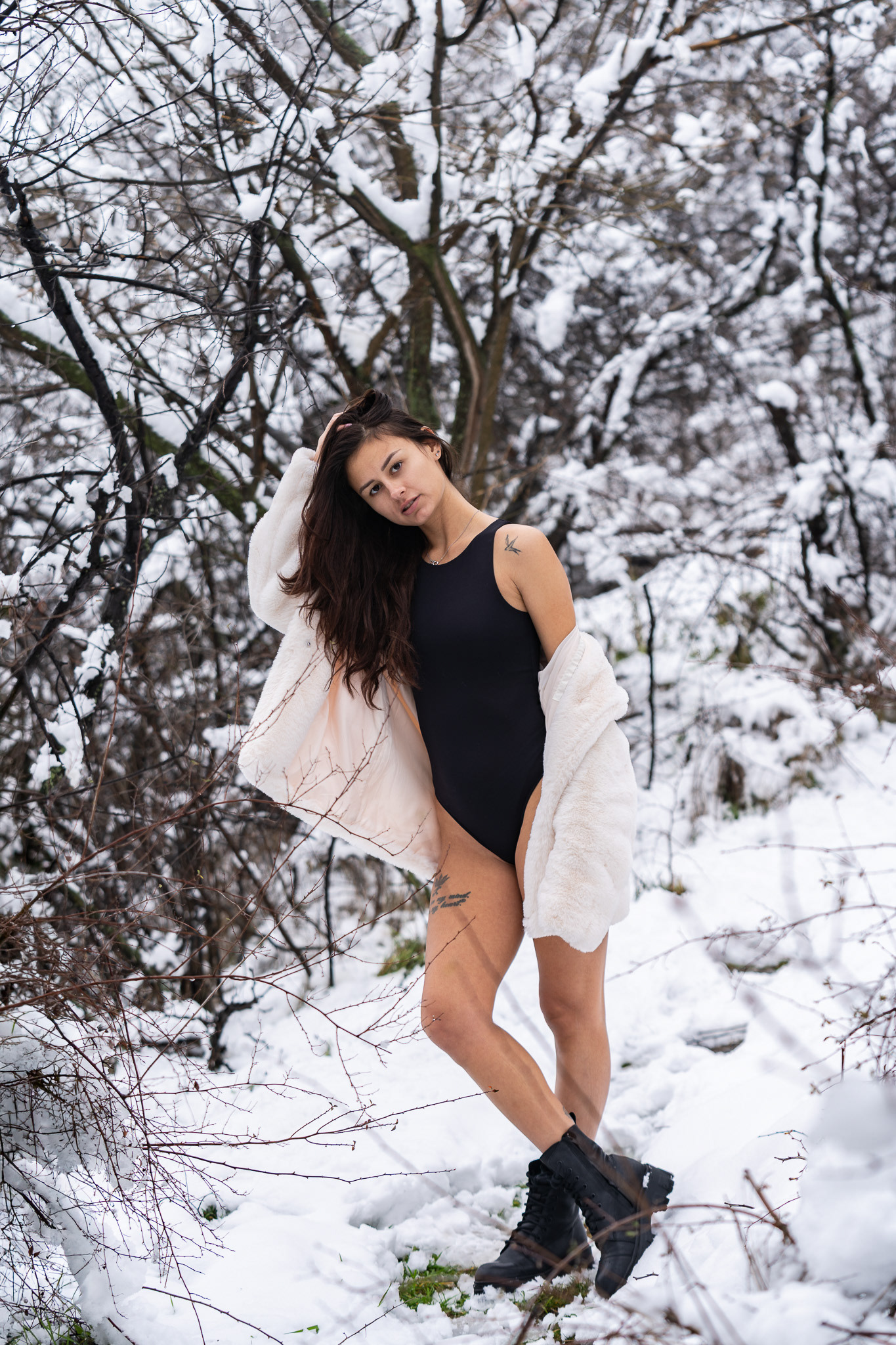 Editorial winter lifestyle shot of a female model in a black bodysuit and open white fur coat with one leg raised among snow-covered woodland. Tenyo Barbolski.
