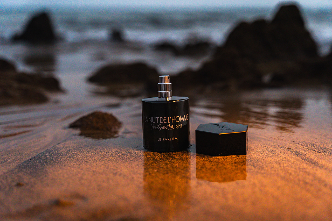 Editorial product landscape of a YSL fragrance on rugged coastal rocks with a mountain silhouette in the background. Focused on global adventure, prestige, and advanced environment compositing.