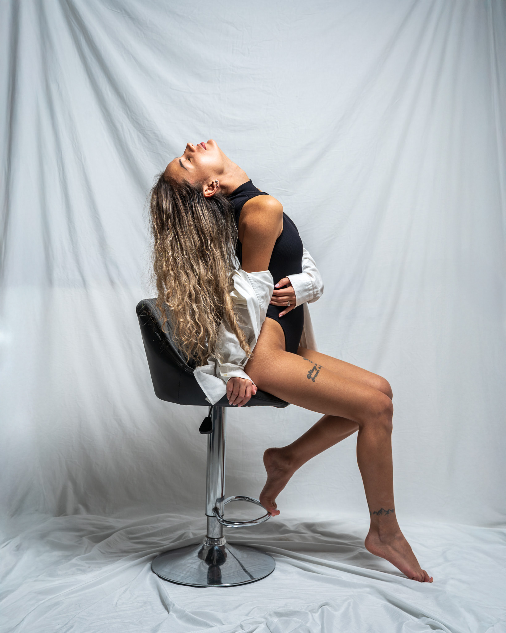 Fine art portrait of a female model arched backward over a chair, utilizing minimalist, high-key lighting. This dynamic pose emphasizes sculptural form, fluid motion, and the clean textures of a white backdrop and black bodysuit. Professional visual storytelling by Arcane Media