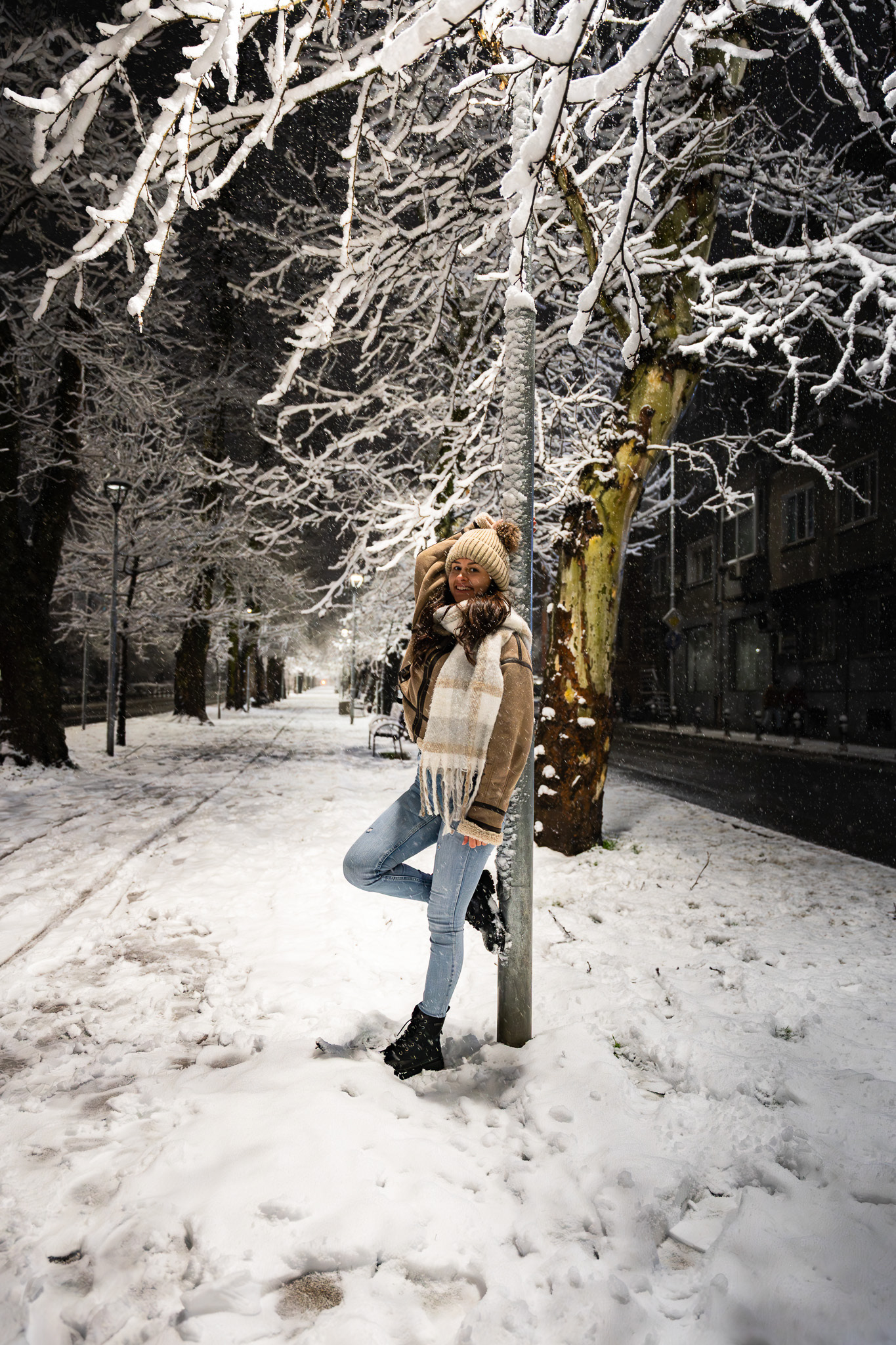 Wide nighttime lifestyle shot of a model in a beanie and winter coat standing beneath a frost-covered lamppost in a snowy park. Editorial photography by Tenyo Barbolski.