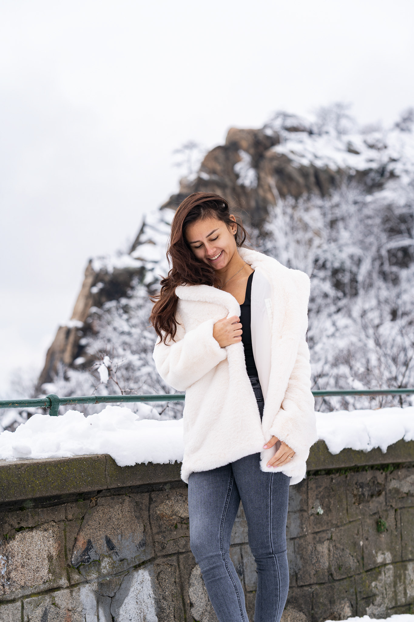 Lifestyle winter portrait of a brunette model in jeans and a white fur coat leaning against a snow-covered stone wall with a rocky hillside backdrop. Tenyo Barbolski.