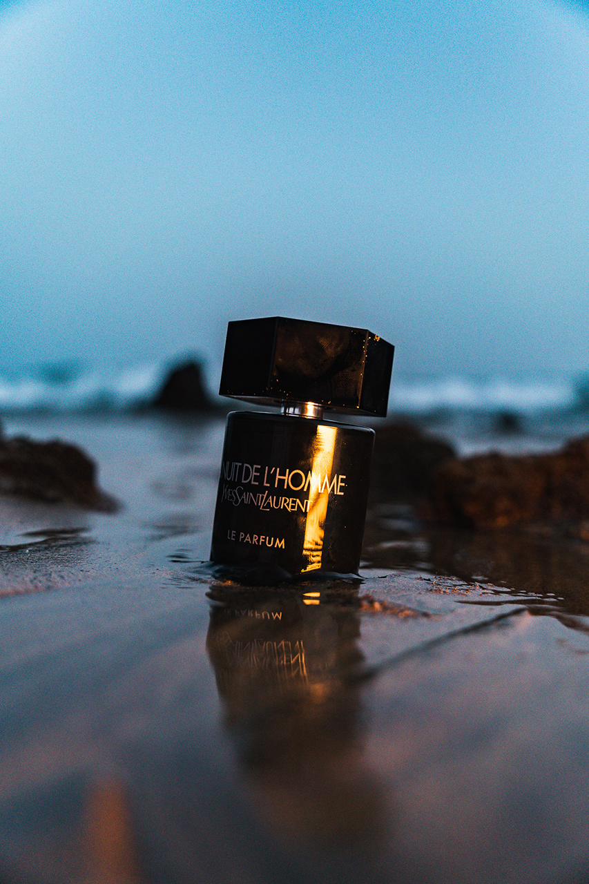 Vertical editorial shot of a designer perfume bottle on a beach, featuring a soft-focus ocean background. High-end lifestyle imagery for luxury fragrance marketing.