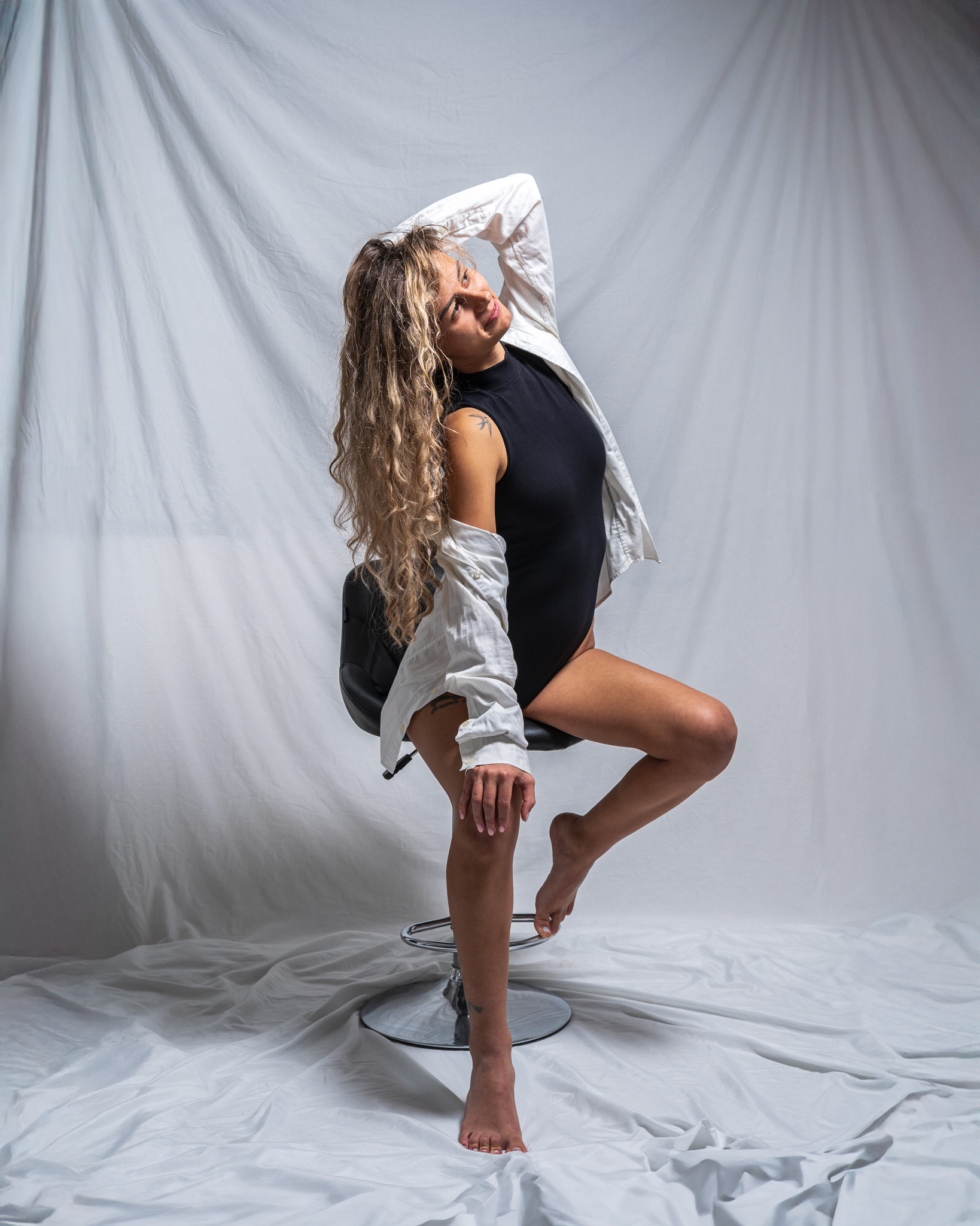 An alternative portrait of a female model in character, using the same studio set (white backdrop/chair). This shot emphasizes a unique, expressive gaze and fluid pose, demonstrating artistic depth in editorial storytelling. Fine art by Arcane Media