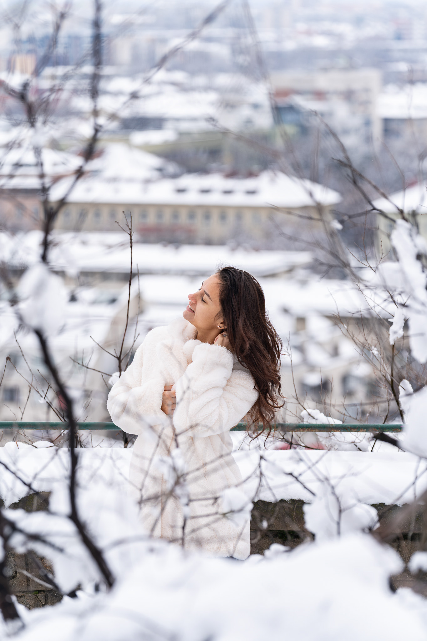 Editorial winter fashion shot of a brunette model in a white fur coat posed against snow-covered rooftops and a mountain in the background. Tenyo Barbolski, Cambridgeshire.