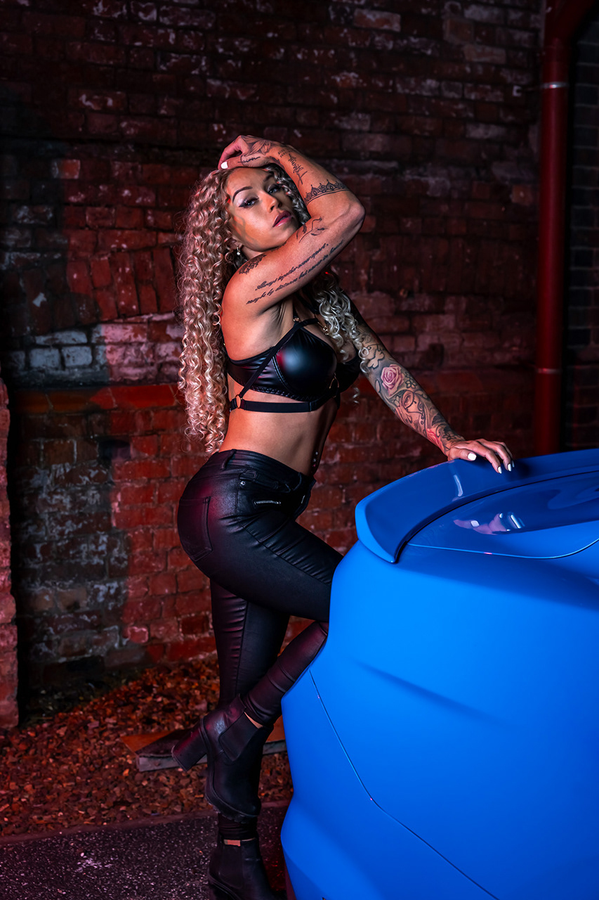 High-contrast urban fashion photography featuring a tattooed gym model in leather styling with a blue Ford Mustang. Expert use of red atmospheric lighting and industrial textures for a dramatic, cinematic editorial look by Arcane Media.