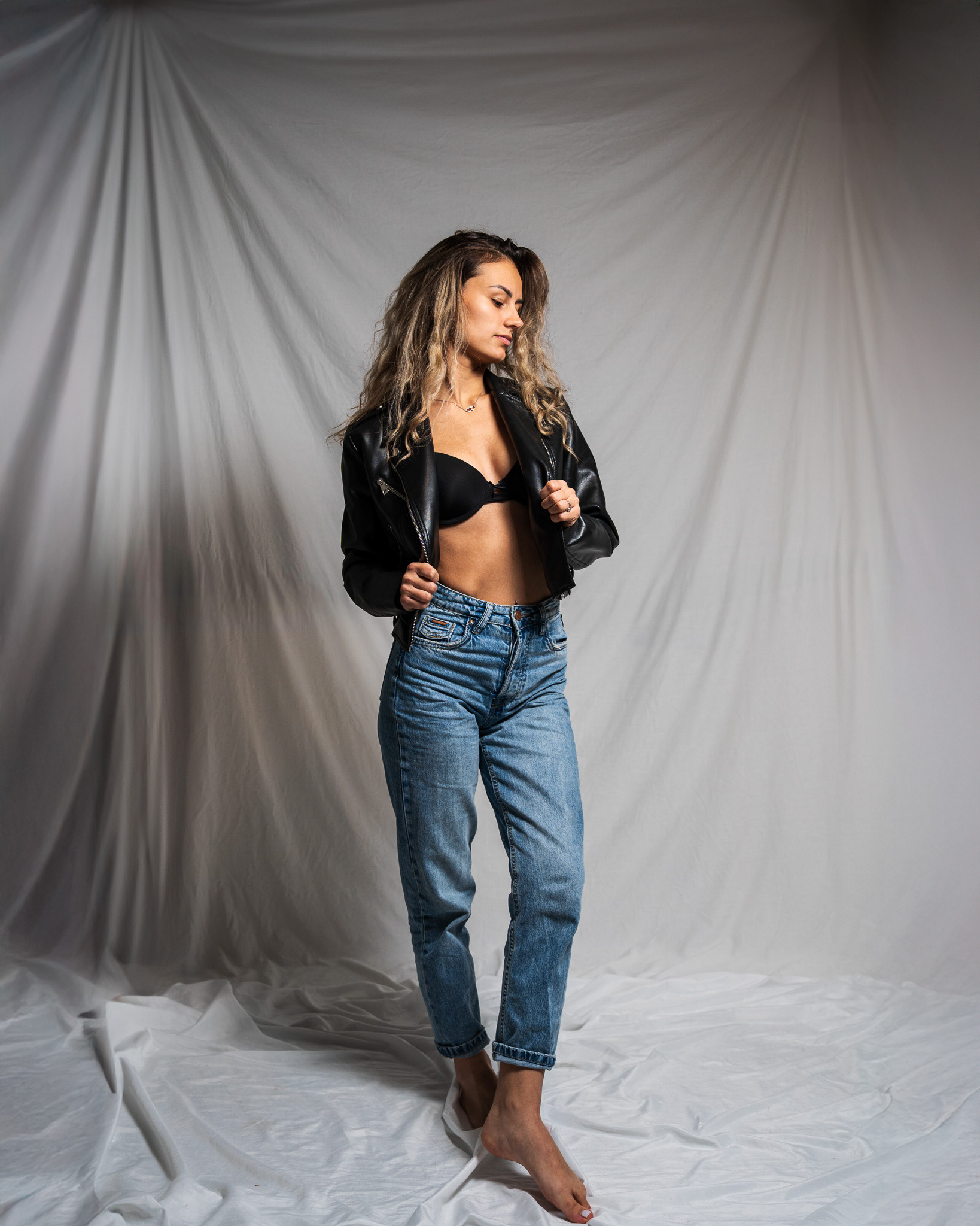 Full-length fashion editorial shot of a model standing in blue jeans and an open leather jacket. Utilizing leading lines from the draped fabric background to create depth and professional scale in a studio environment. Tenyo Barbolski.