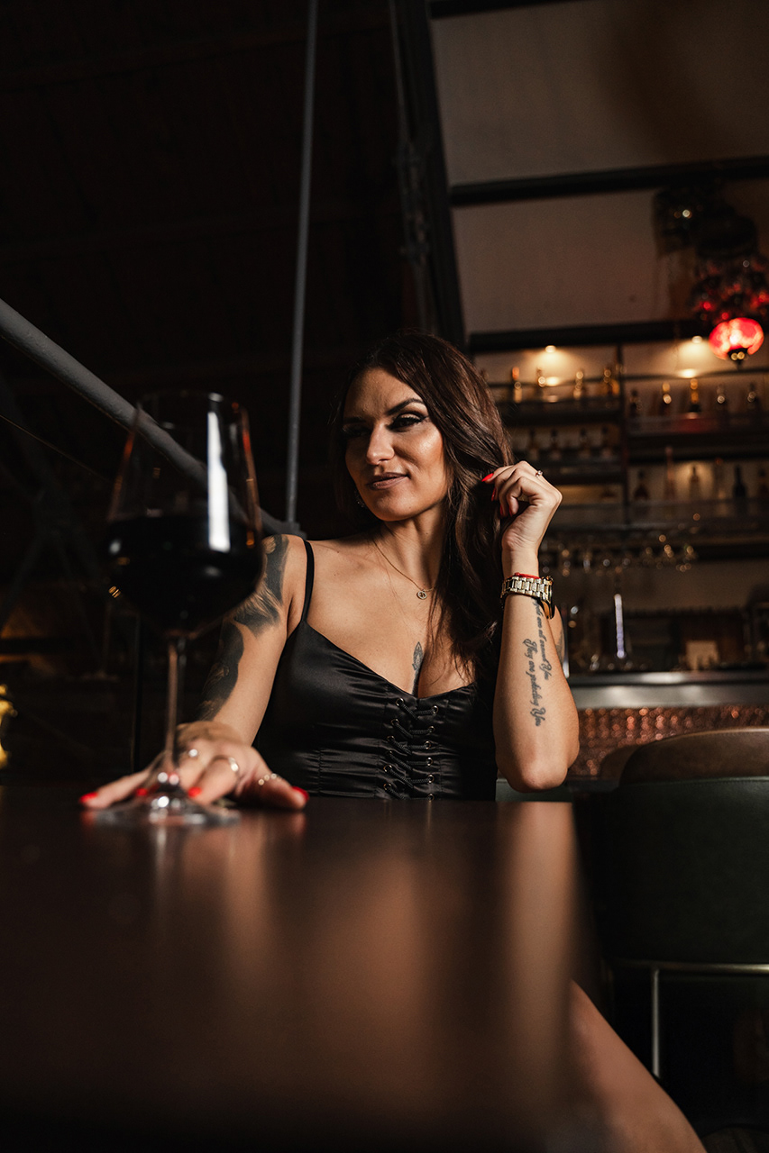 Close-up editorial portrait in a dark bar setting. Atmospheric lighting focuses on the model’s face and the high-end mood of the location.