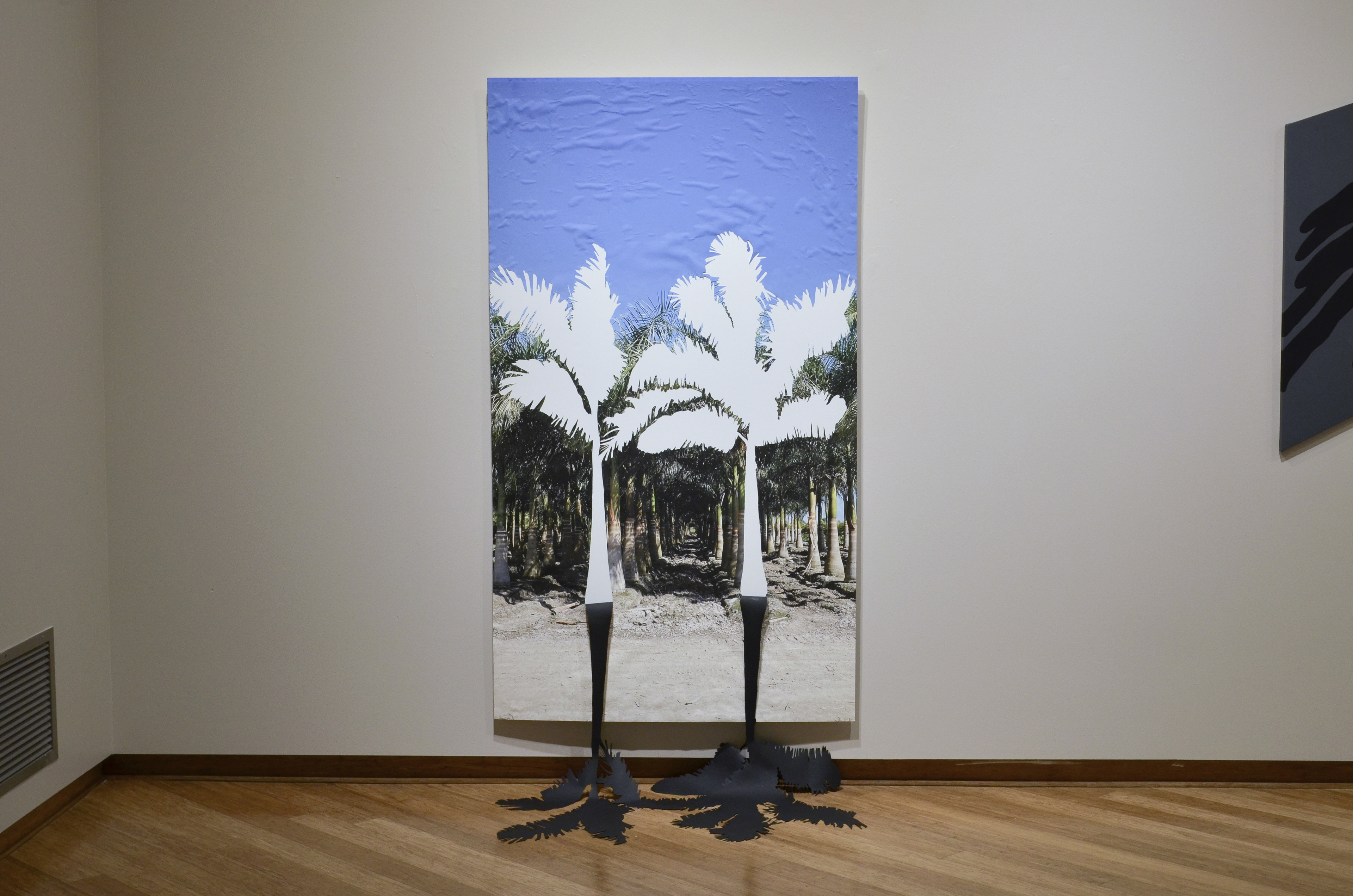 A large-format image of a palm tree plantation in a gallery. The first row of trees has been cut out, and is falling on the floor, creating a black, physical shadow in the gallery.