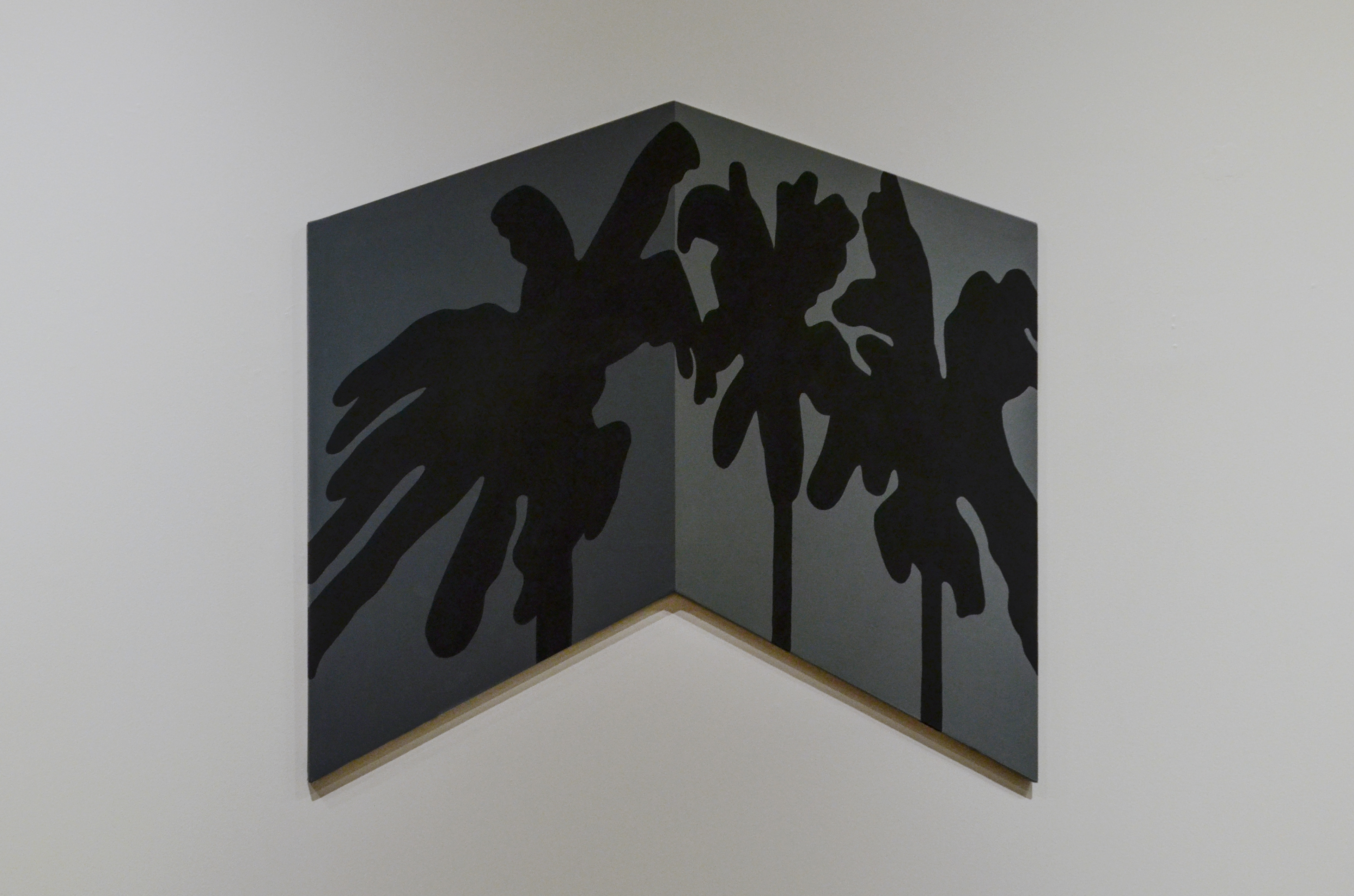 A shaped canvas depicting three palm trees casting shadows along the corner of a wall. The painting appears to be 3-dimensional, but exists on a flat surface in the shape of a chevron.