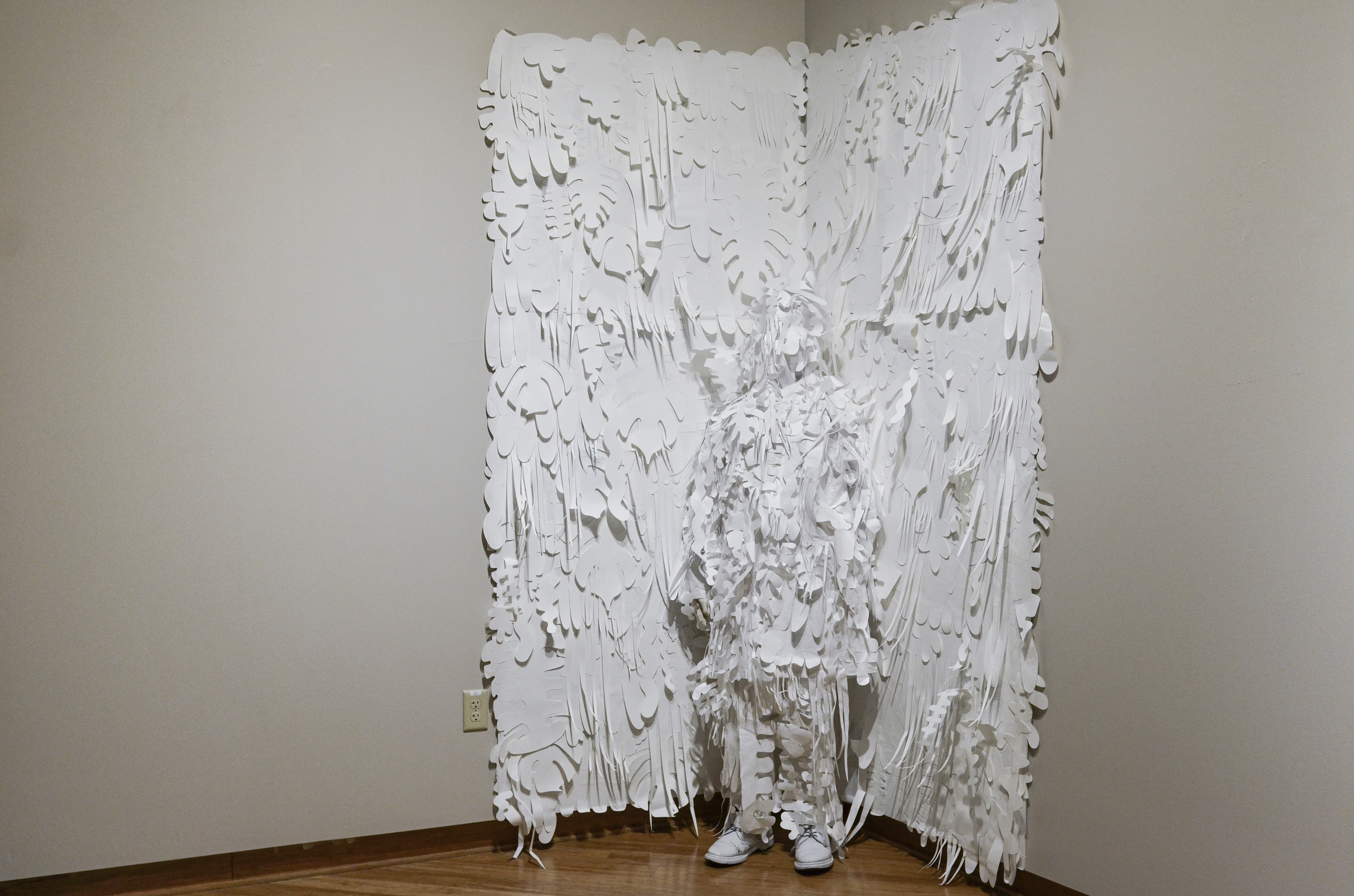 A camouflaged figure blends into a wall covered in textured and abstracted white tropic foliage.