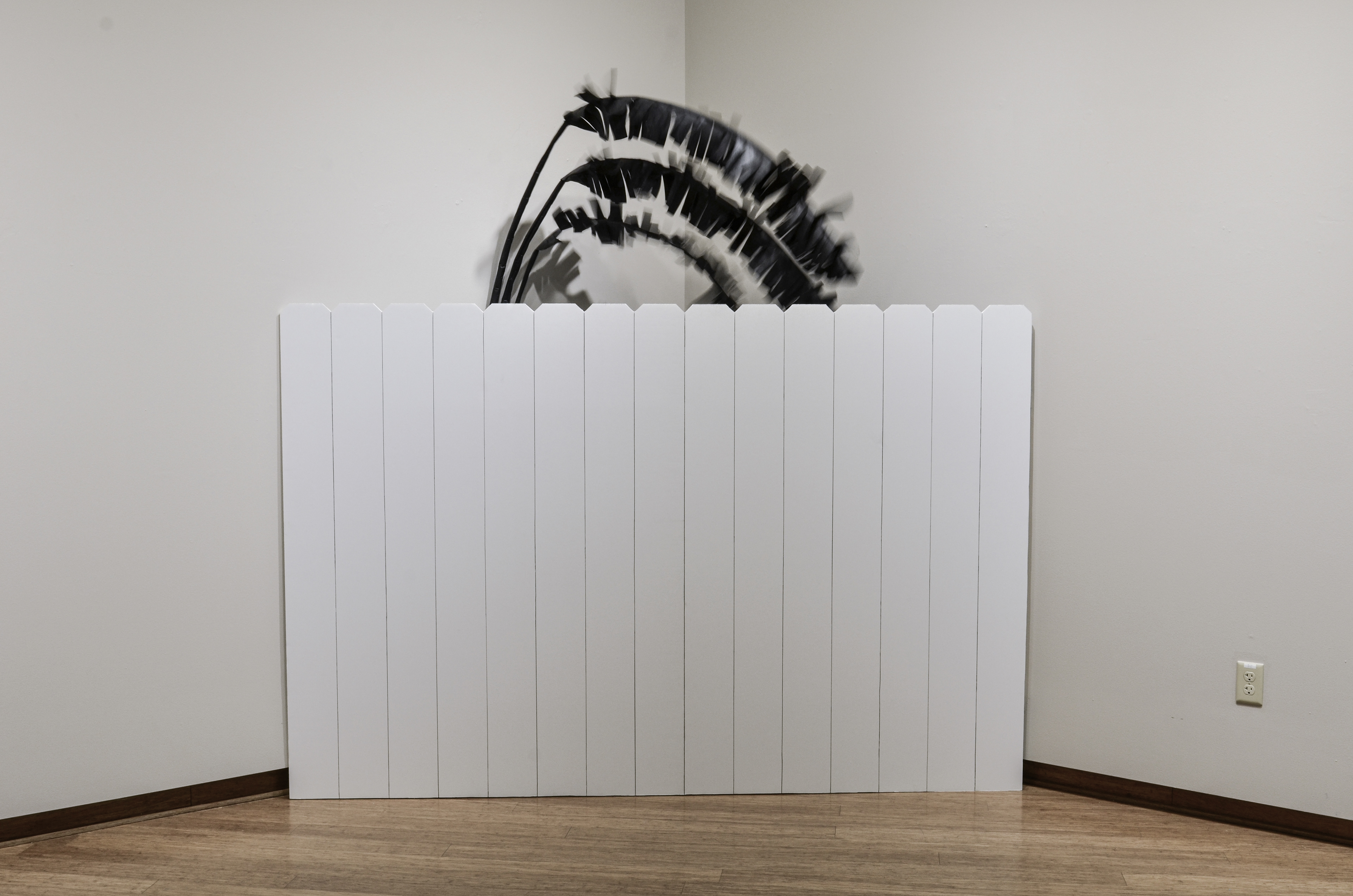 A time-lapse photo of a kinetic sculpture of a black banana plant behind a fence, resting in the corner of the gallery. The fan (hidden behind the fence) agitates the leaves.